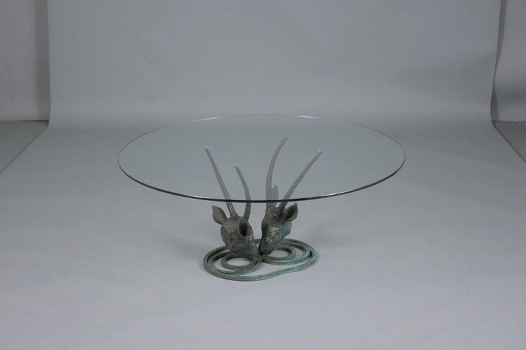 An extraordinary mid-century modern ibex coffee table hand-crafted out of bronze and newly restored by our team of craftsmen. This piece features an eye-catching ibex rams head design base with a unique patina finish supporting a round glass top