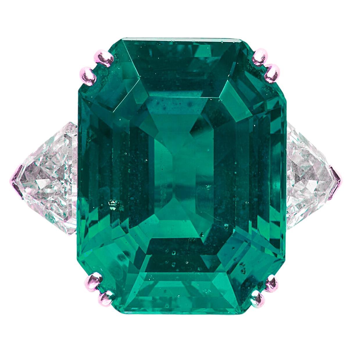 This exquisite ring features a ICA Certified 12.89 carat vivid green emerald as its stunning centerpiece, radiating a rich and captivating hue. Flanking the emerald are two shield-cut diamonds, adding a touch of brilliance and enhancing the ring's