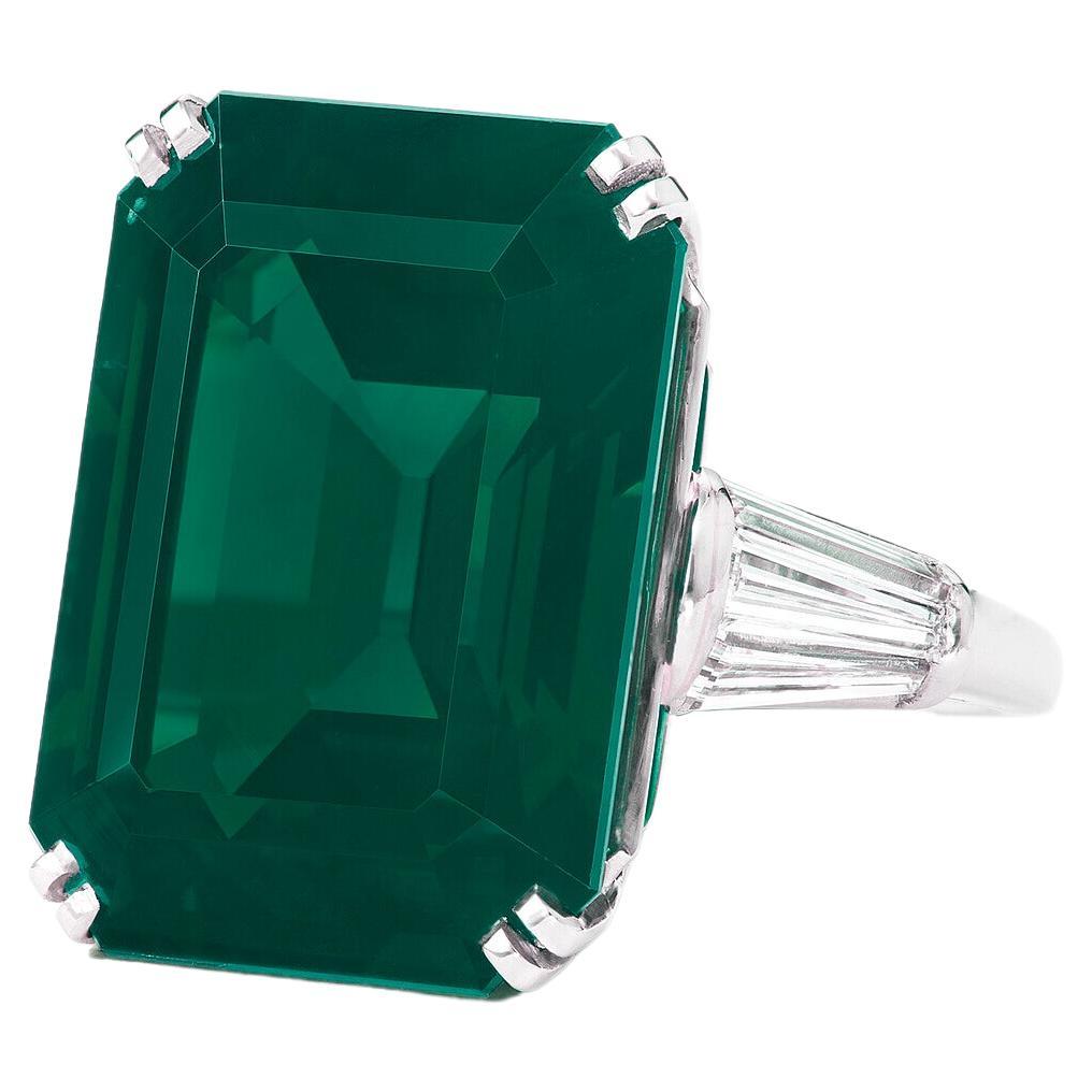 This exquisite ring features a ICA Certified 15 carat vivid green emerald as its stunning centerpiece, radiating a rich and captivating hue. Flanking the emerald are two shield-cut diamonds, adding a touch of brilliance and enhancing the ring's