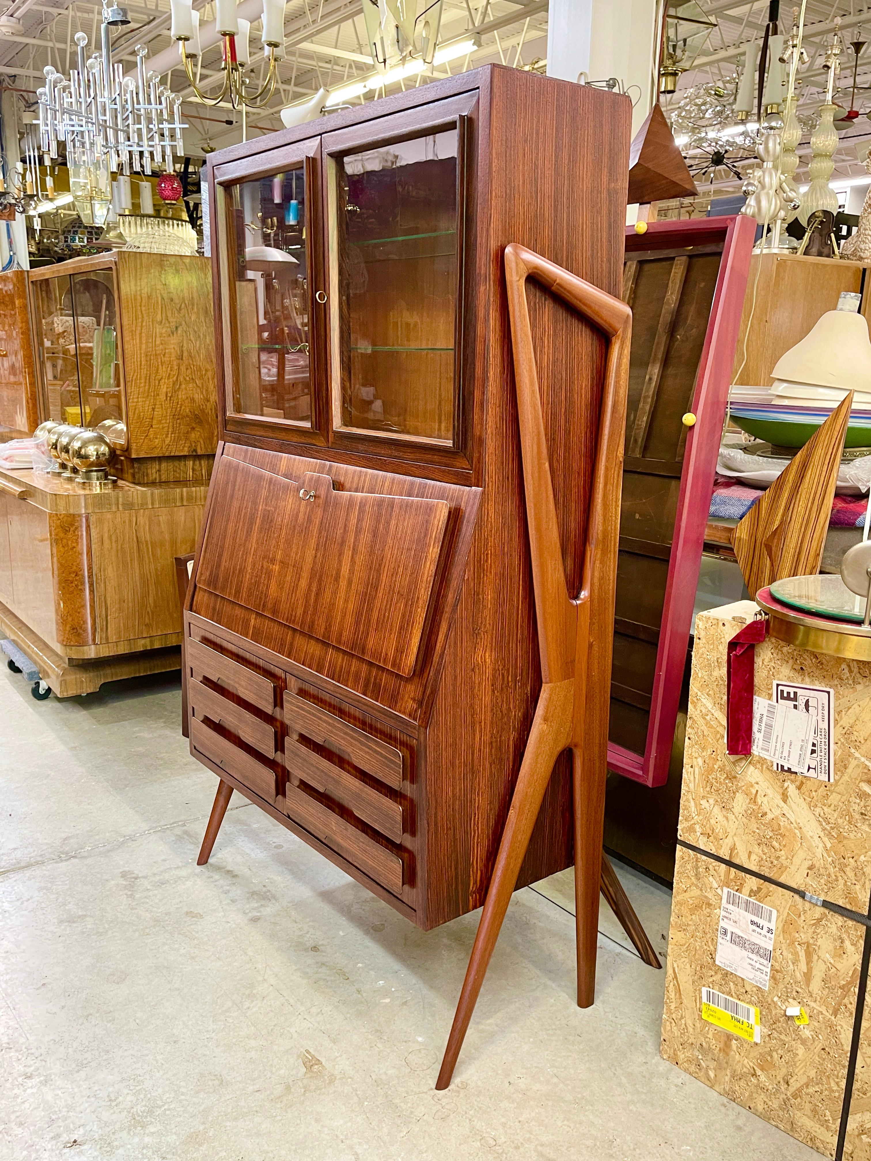 1950s Italian liquor bar cabinet or secretary writing desk book cabinet in rosewood with distinctive sculptural ebonized and scaffold-like legs. 
Pair of glass doors open to reveal two adjustable glass shelves. 
Drop front door with brass key.