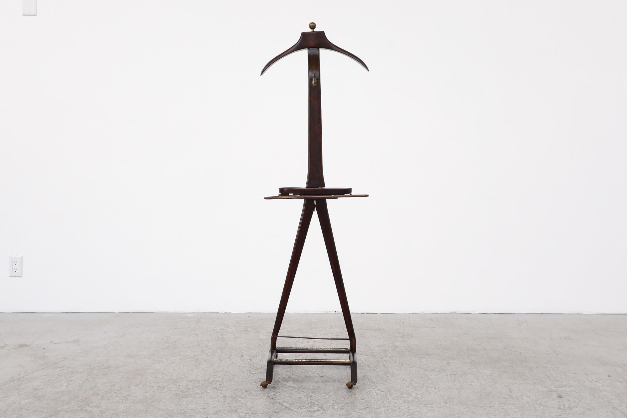 This Italian sculpted rolling clothing valet stand is designed by Ico Parisi, 1960, Italy. It features a molded wood cufflink and jewelry tray, pull out tie bars, jacket and pant holder, a brass hook and a shoe stand. It is in very original