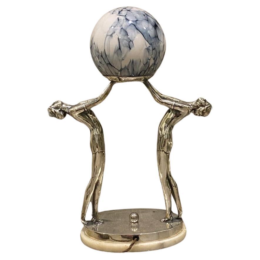 Superb Iconic Twin Lady Art Deco Lady Table/Desk Lamp
A superb, scarce and stylish Art Deco table or desk lamp of two dancing ladies in silvered spelter holding aloft a Loetz type blue veined glass globe mounted on a rectangular alabaster base.
The