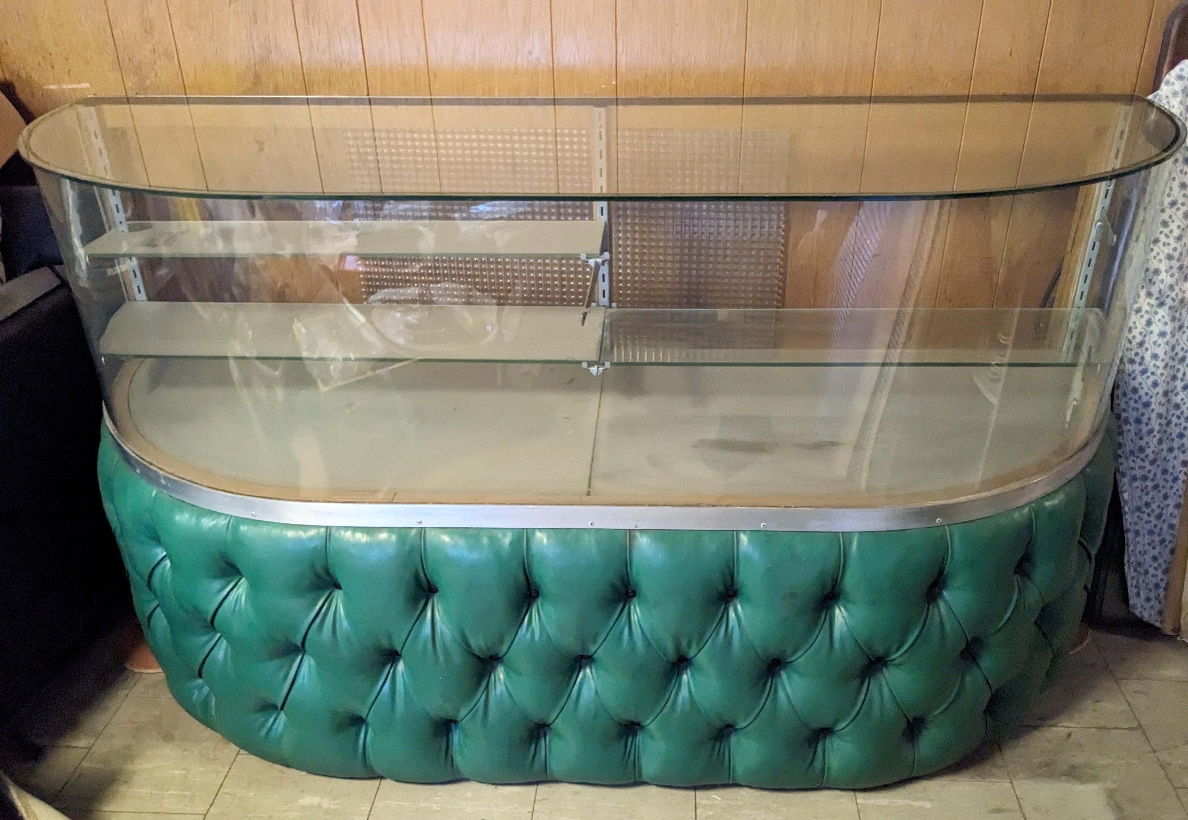 Iconic Mid Century Curved Vitrine Showcase with tufted Naugahyde base circa 1940-1950 of American origin. Vitrine has storage behind/below the 1950's green tufted vinyl with its original waffle glass sliding doors.
Showcase also has a switch to