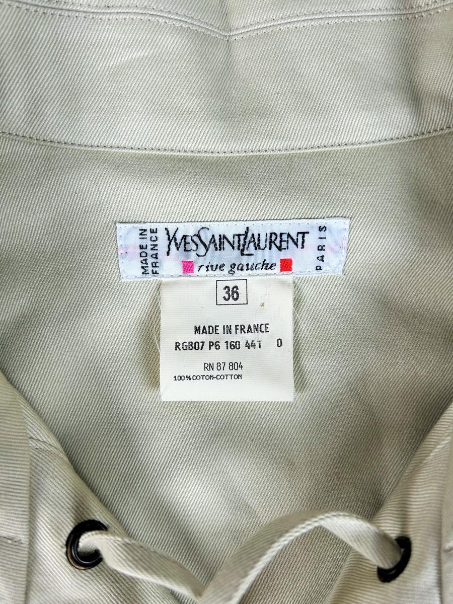 Circa 1995 - France
Iconic Yves Saint Laurent Rive Gauche blouse jacket in grey twill cotton gabardine dating from the 1990s. Quite similar to the 1969 Saharienne with its vertical lacing with metal rivets and four real flap pockets adorned with