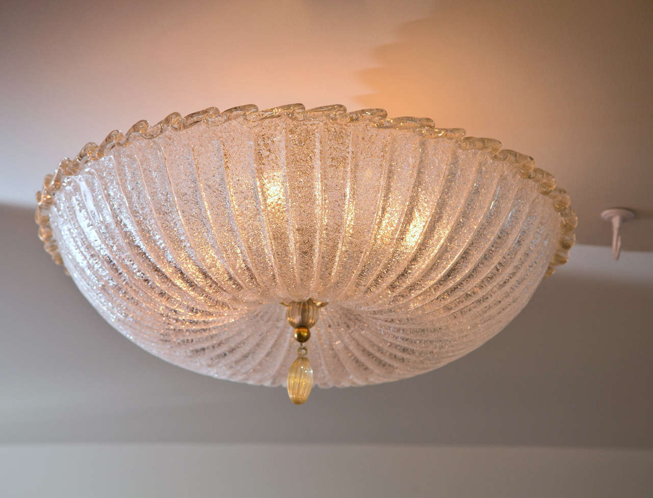 Large shimmery pin cushion shaped ceiling fixture with a lightly gold blown rim, disk and tassel. When illuminated it takes on a beautiful icy effect.
This fixture will be made install ready with unlacquered brass hardware to match the brass detail