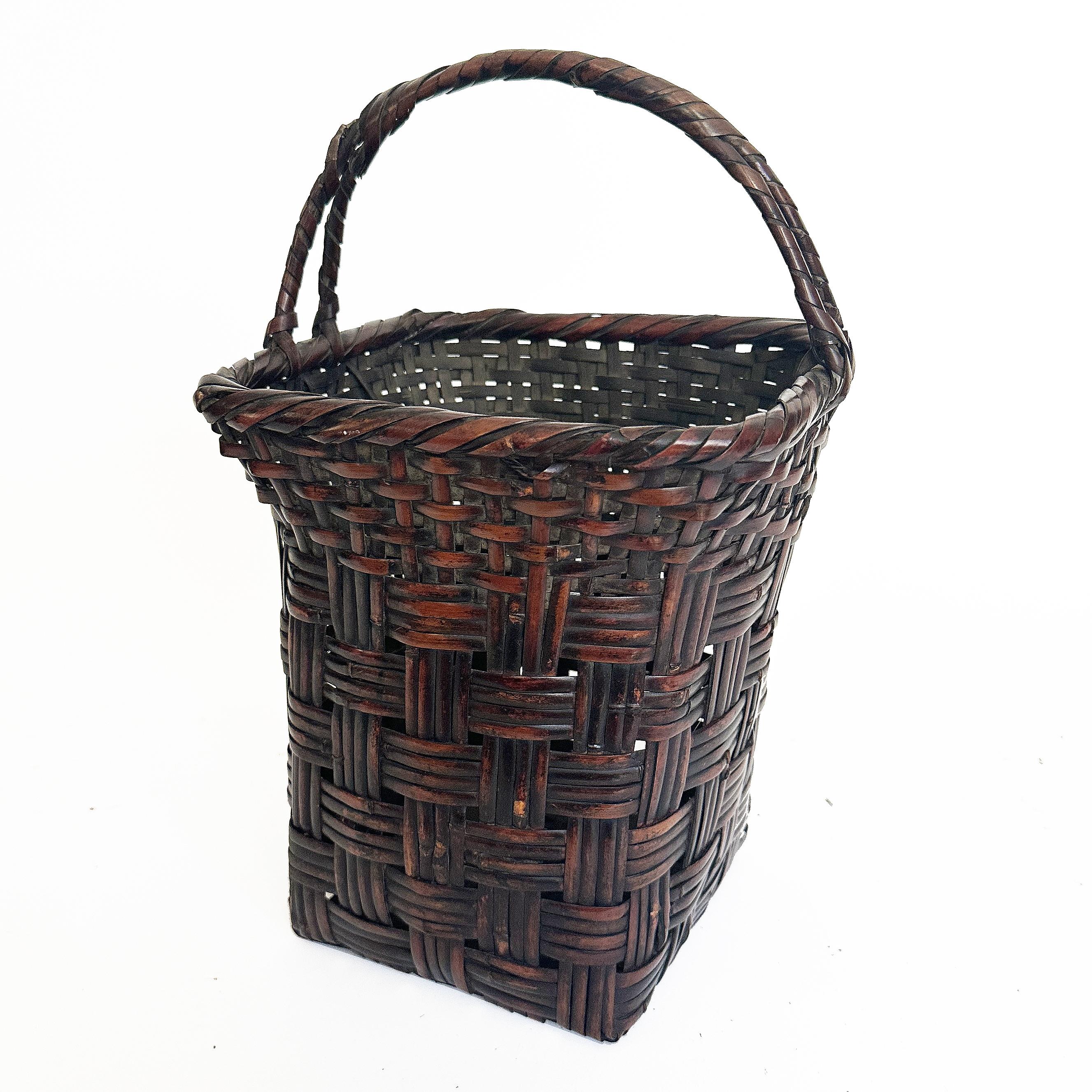 This ikebana basket features two weave patterns with a twisted handle in a square shape. It has a deep brown color with a slight contrast to highlight the weaving. It is constructed from bamboo strips of various sizes. Ikebana baskets are closely