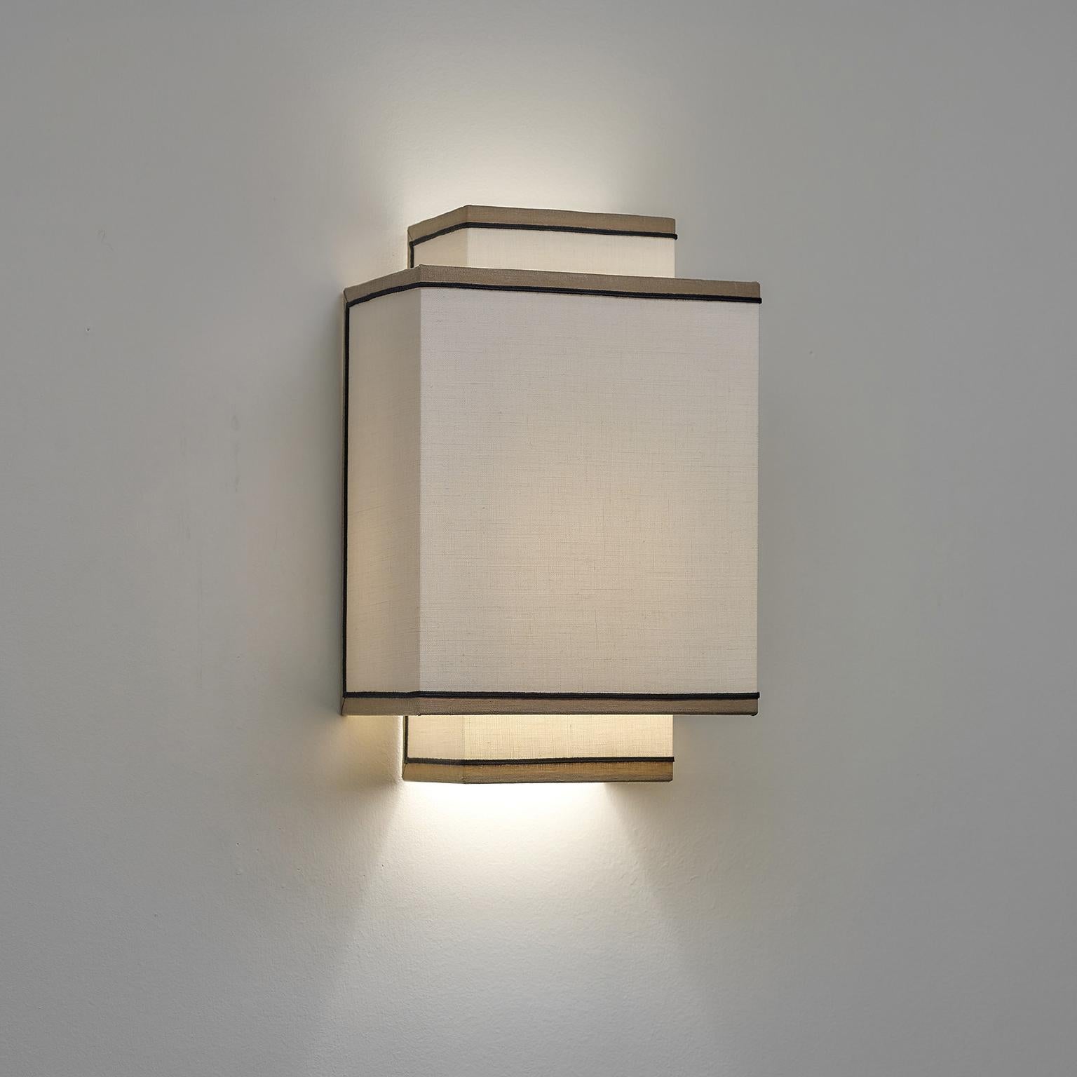 This striking wall sconce is a testament to architectural lines and elegant simplicity. This sconce is characterized by a geometric silhouette composed of two intersecting rectangles handcrafted of ivory linen with subtle taupe rim and black