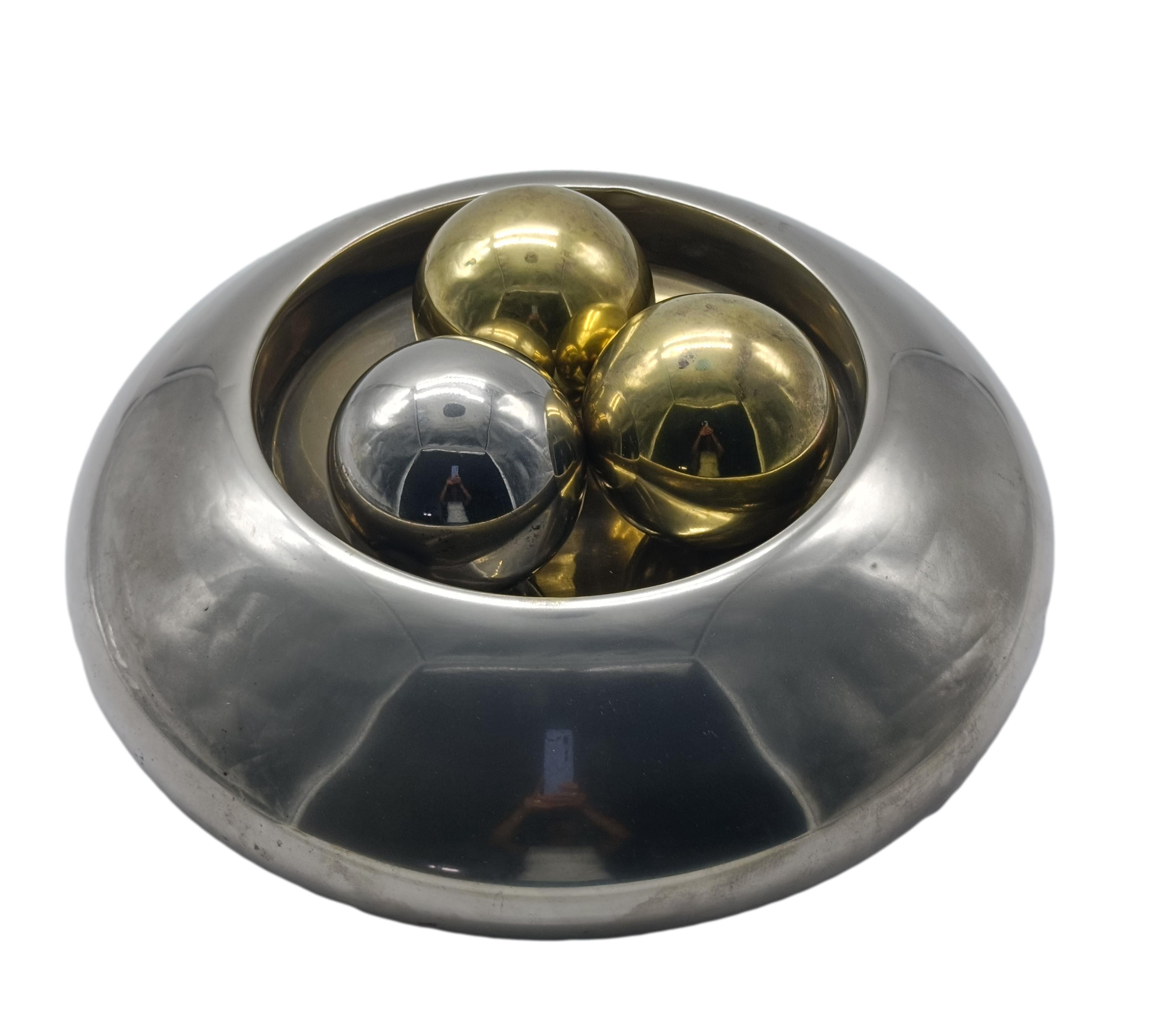 Elegante y original centro de mesa de los años 70, de acero inoxidable pulido con inserciones esféricas de latón dorado y acero cromado. La estructura principal tiene una forma redondeada y cóncava, casi parecida a un donut o anillo elíptico, que