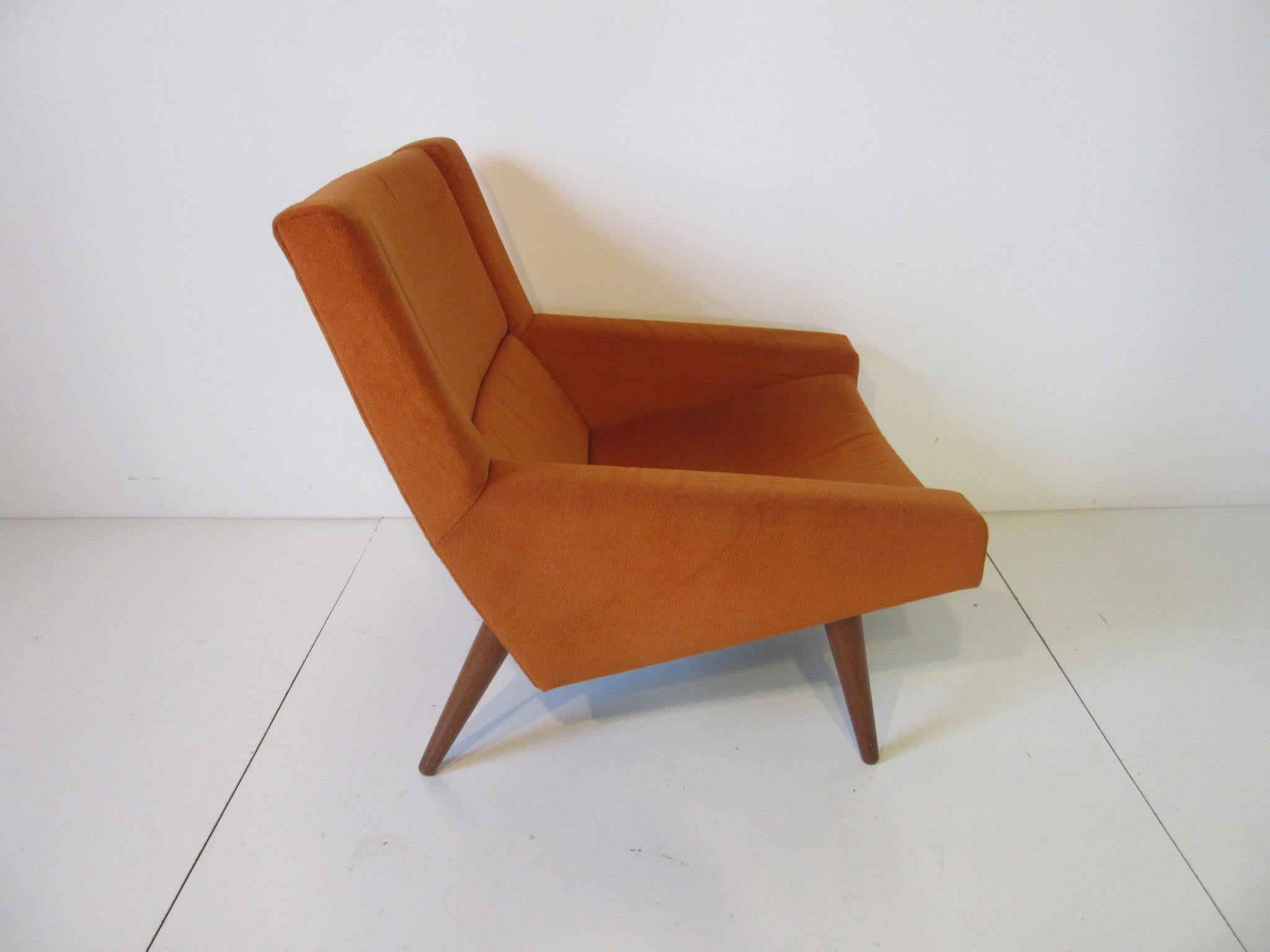 A well crafted upholstered lounge chair in a soft orange fabric with conical solid teak wood legs and having an interesting ergonomically designed shape for comfort. Purchased by the original owners in Denmark and lovingly cared for until their