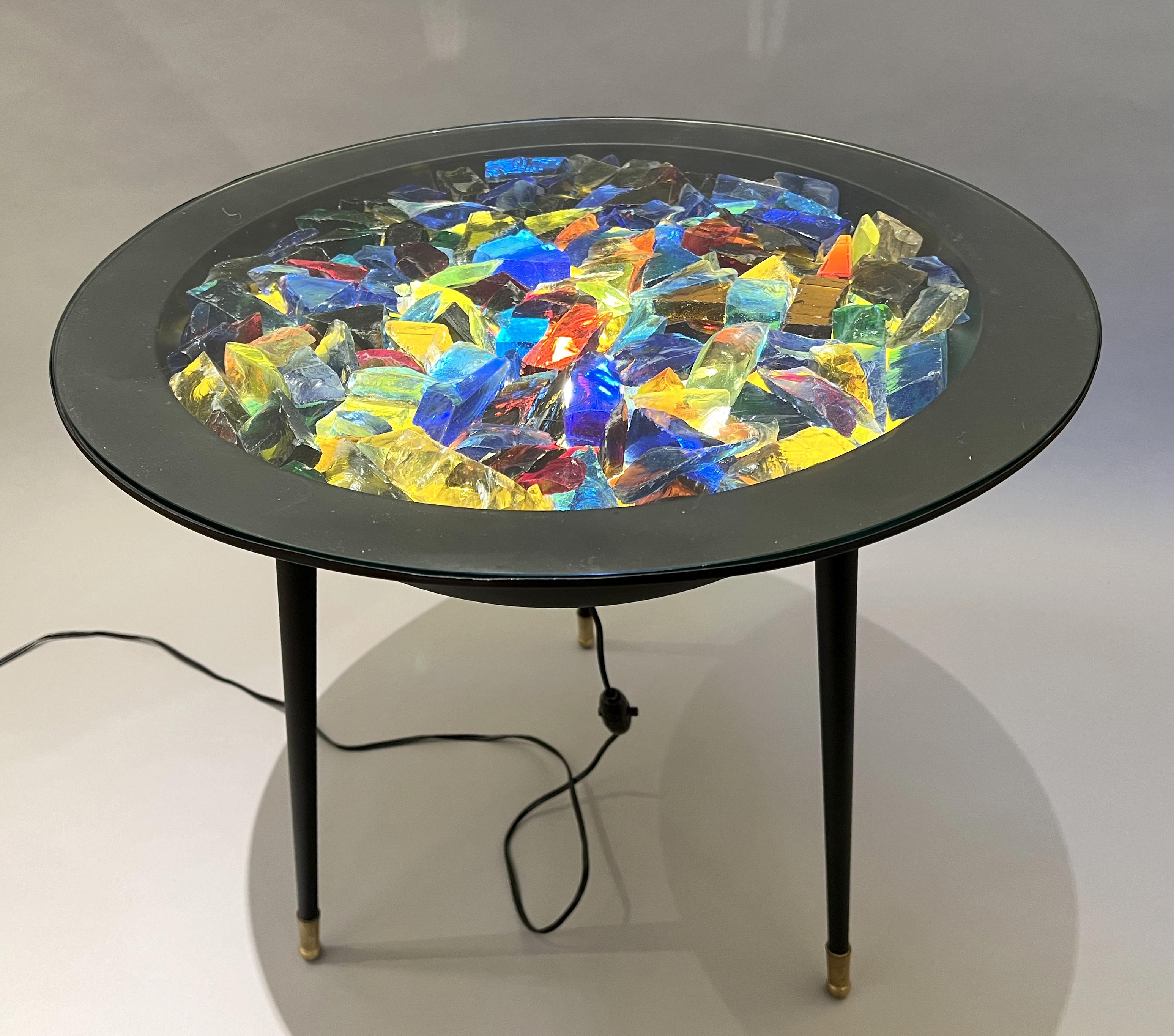 Interesting luminous pedestal table featuring a round basin with three black-lacquered iron legs. The legs are finished with brass sabots.
The basin contains multicolored glass pieces trapped between two glass plates. It is lit from below by an