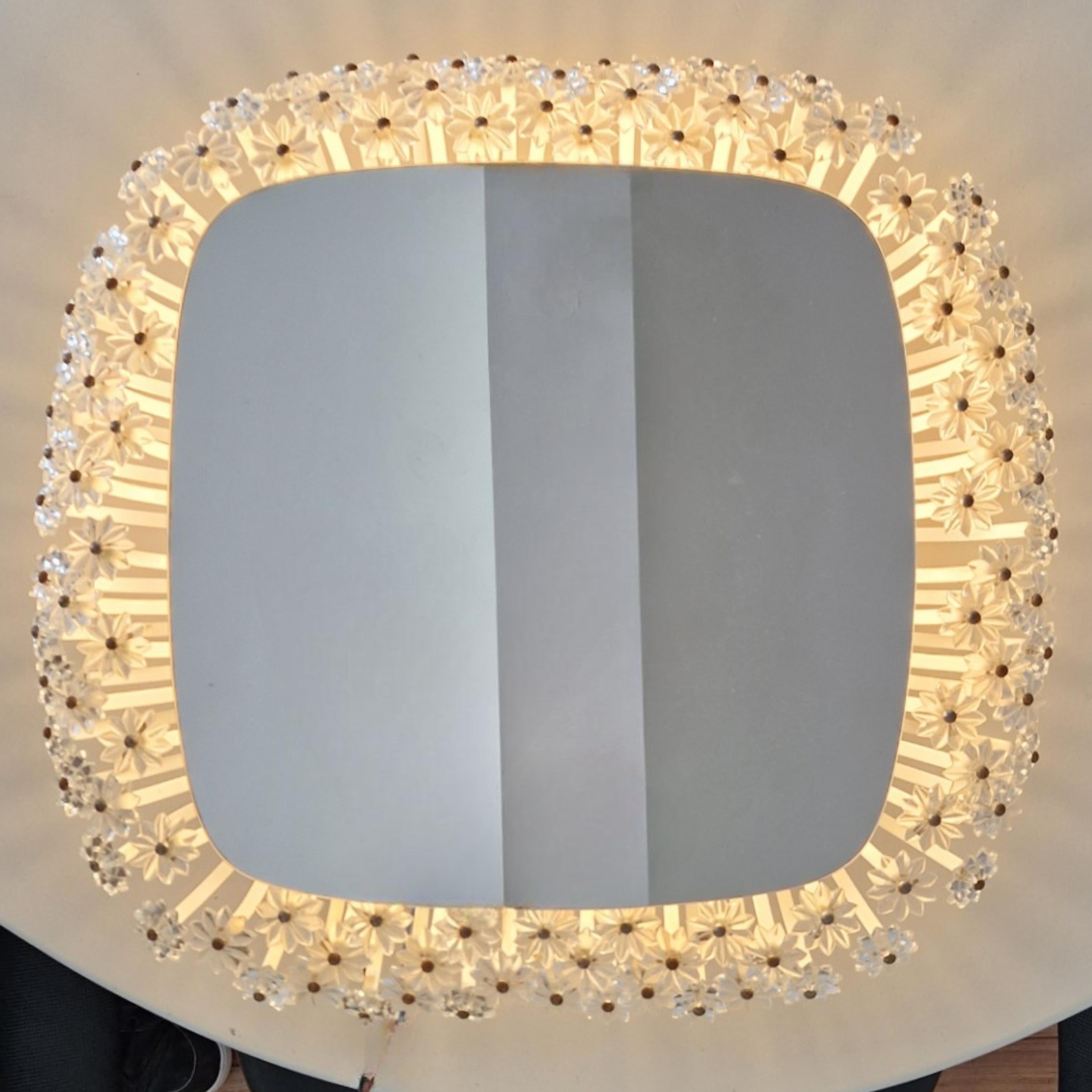 Back lit wall mirror. Designed by Emil Stejnar and produced by Rupert Nikoll, Austria in the 1950s. The mirror has an edge made from numerous glass flowers that reflect en disperse the light coming from behind the mirror. It has one E27 European