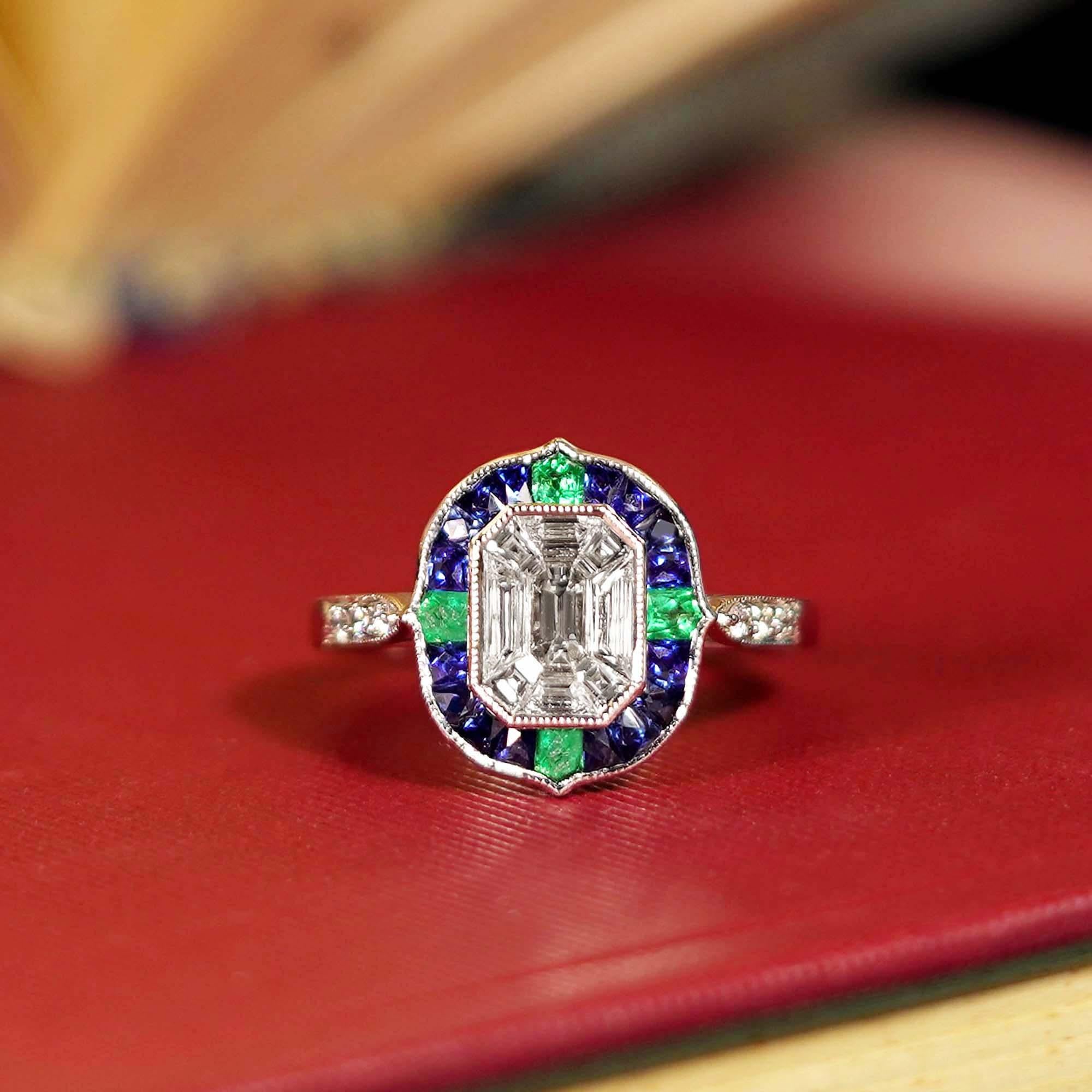 A stunning illusion setting diamond at the center, creating a bold and captivating sparkle. The diamond is surrounded by rich sapphires, adding depth and vibrant contrast. Delicate emerald accents decorate the top, left, right, and bottom, enhancing
