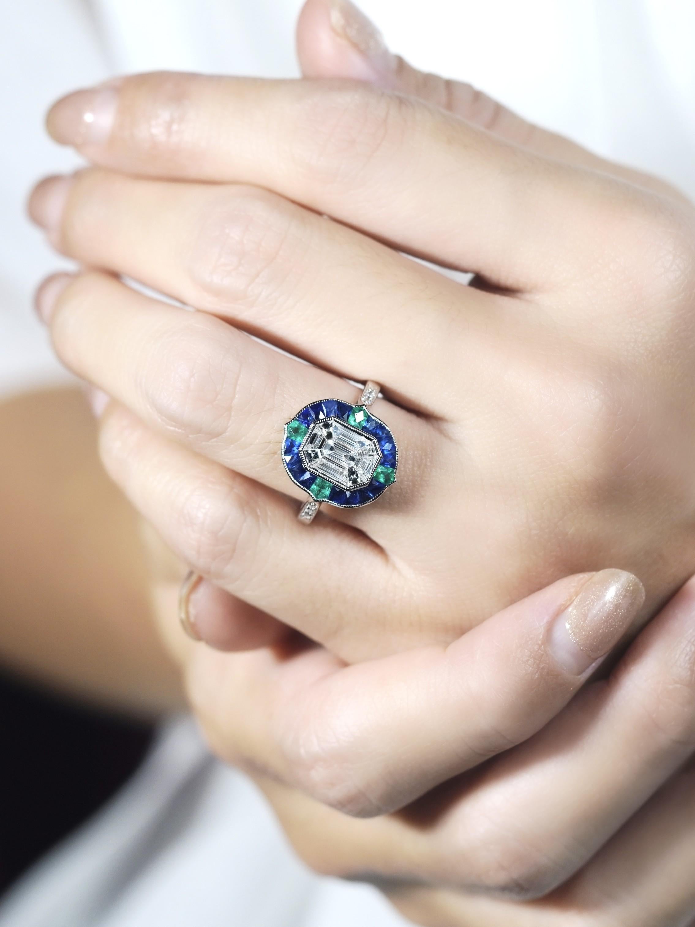 A stunning illusion setting diamond at the center, creating a bold and captivating sparkle. The diamond is surrounded by rich sapphires, adding depth and vibrant contrast. Delicate emerald accents decorate the top, left, right, and bottom, enhancing
