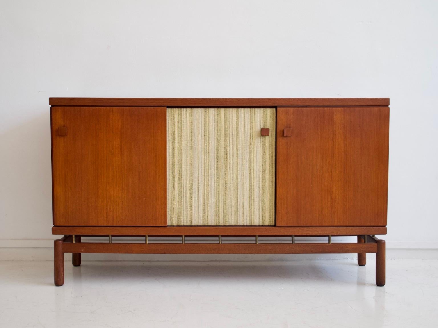 Sideboard made of teak wood with brass details. Sliding doors, one of them decorated with fabric. Produced by La Permanente Mobili Cantù and designed by Finnish interior architect Ilmari Tapiovaara.

Literature: Selettiva Cantù Catalog.