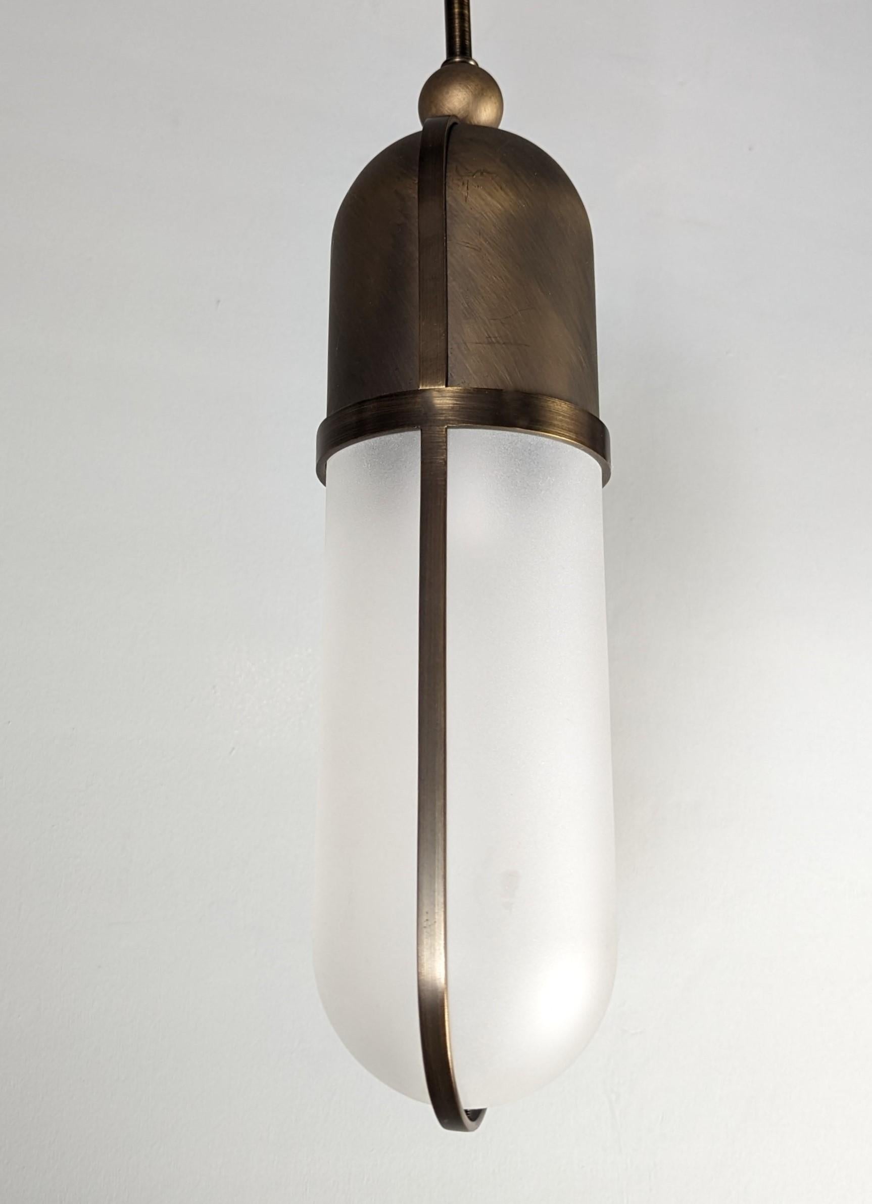 Inspiration for this pendant lamp is taken from “The Century Building” Lobby in New York. This collection is crafted from vertical rounded linear bronze frame with glass and brass shade insert to either sides. Sleek and elegant antique bronze and