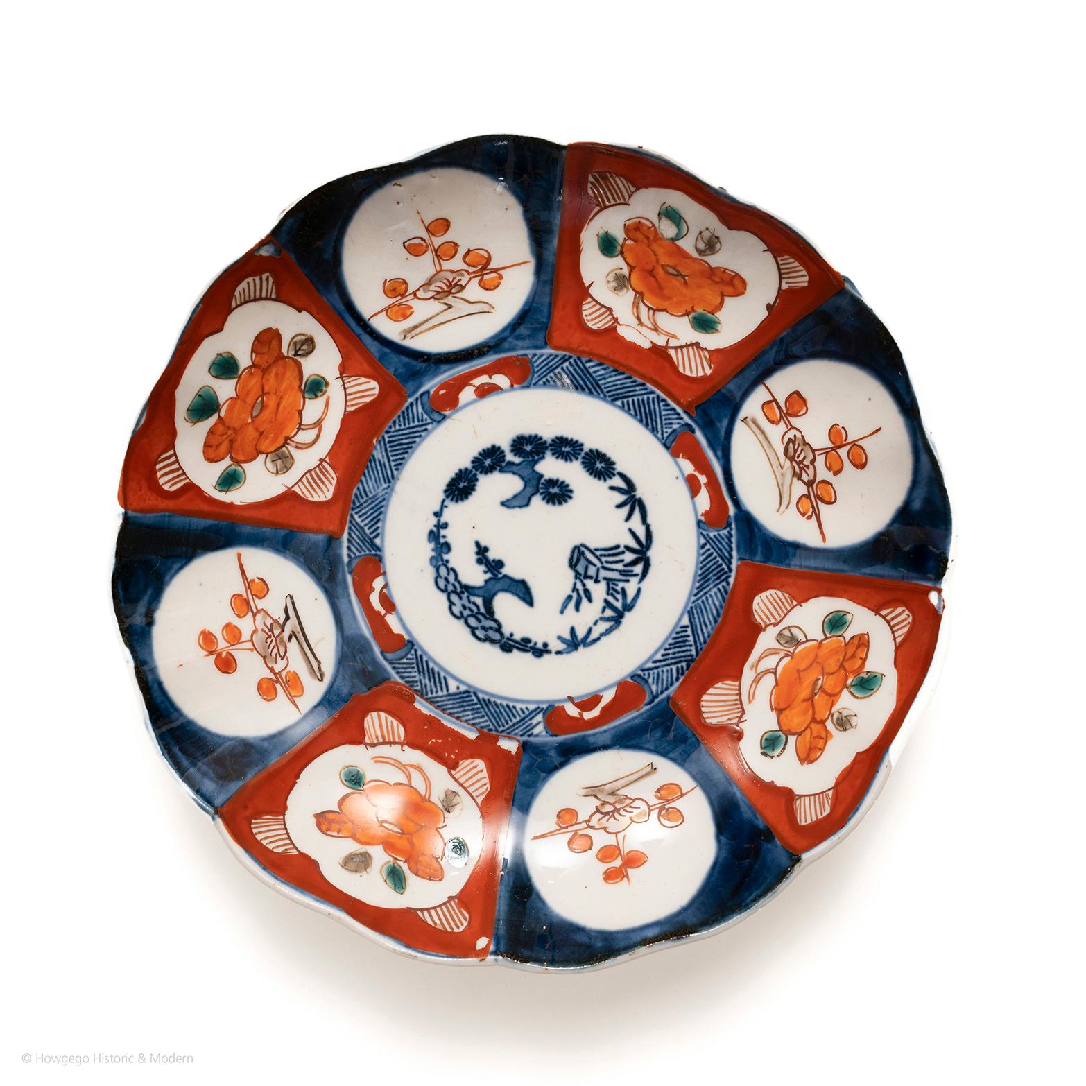 Auffälliger, gelappter Imari-Teller mit einfachen, kühnen Ornamenten, die auf ein frühes Stück hindeuten. In der Mitte drei stilisierte Bonsai, die an die Qualität der Reflexion erinnern, umgeben von einem Spalier und einer Blumenrabatte. Der Rand