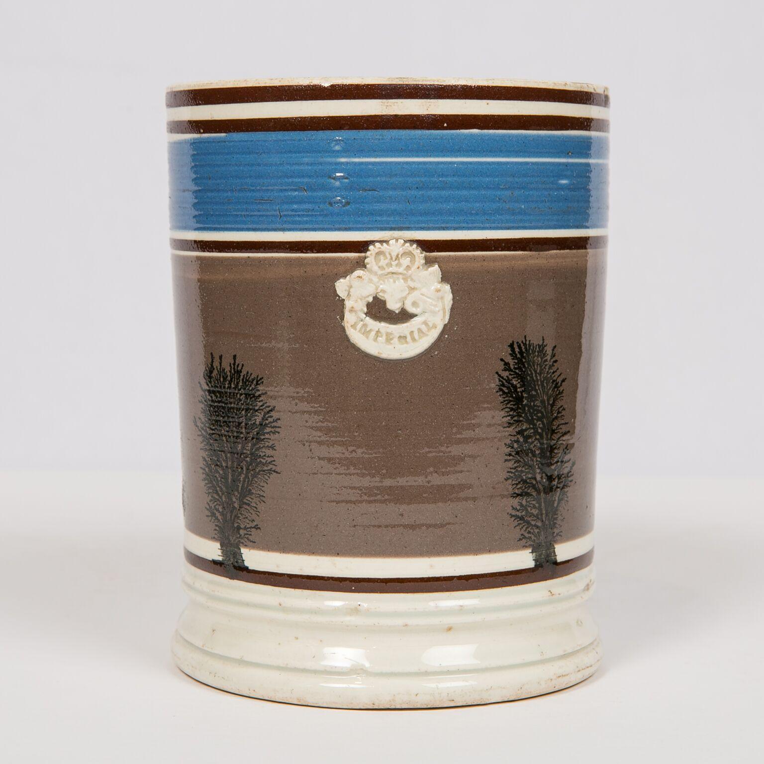 T.his is an Imperial Quart mocha ware mug. The mug was made in England in a style that was popular on the European Continent. It has rouletted bands covered in a blue slip and a large field of gray-brown slip that is decorated with 