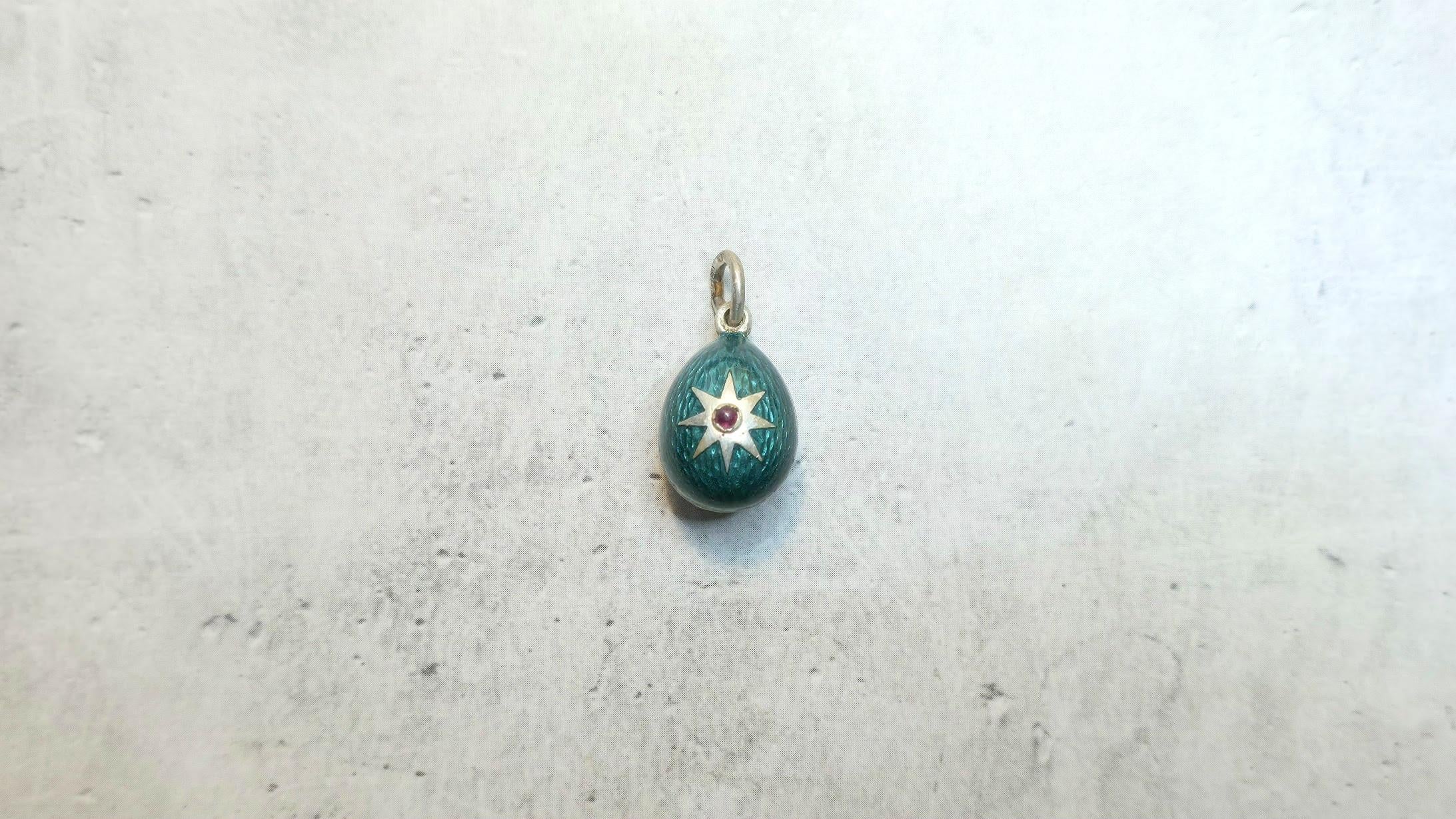 Questo splendido ciondolo a forma di uovo, realizzato in argento dorato, presenta una vibrante finitura in smalto verde guilloché. È elegantemente ornato da un motivo a stella su un lato, accentato da un piccolo rubino naturale: un pezzo davvero
