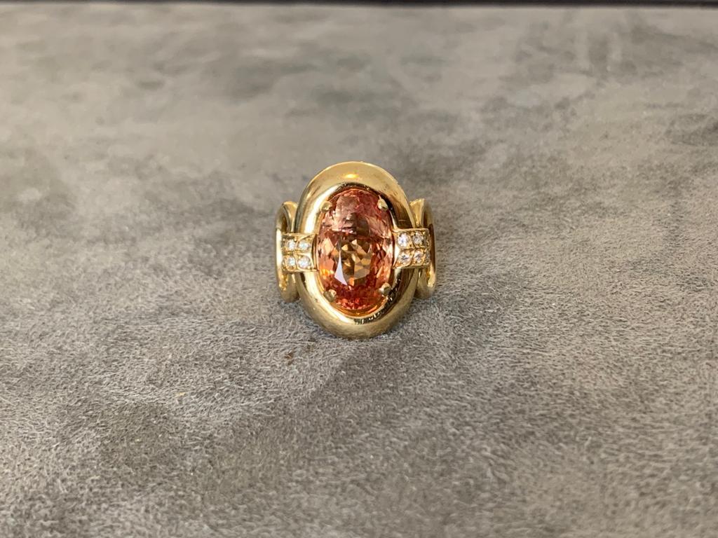 An orange Imperial Topaz of approximately 7 ct (stone's dimensions 15.1x9.3x7.2mm) set in 18k yellow gold. 8 diamonds of about 1mm set on each side of the ring. The Topaz has a large visible crack across the table, and the shape of the shank is