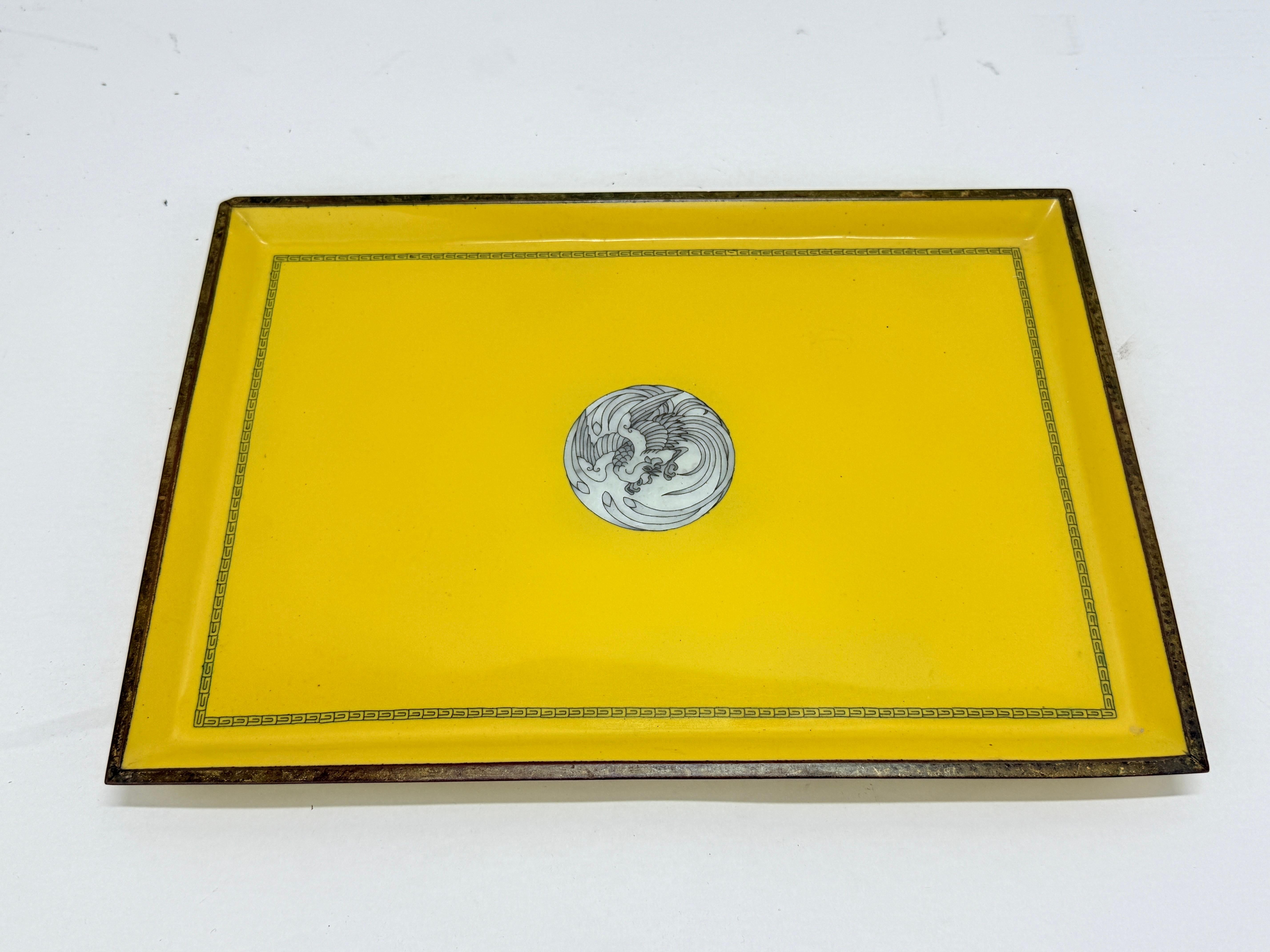 Raffinato vassoio rettangolare in smalto cloisonné su rame, il cui campo è rifinito in un giallo imperiale saturo e centrato da un tondo con un hō-ō (fenice) stilizzato tra nuvole vorticose. La composizione è tranquillamente formale: una sottile