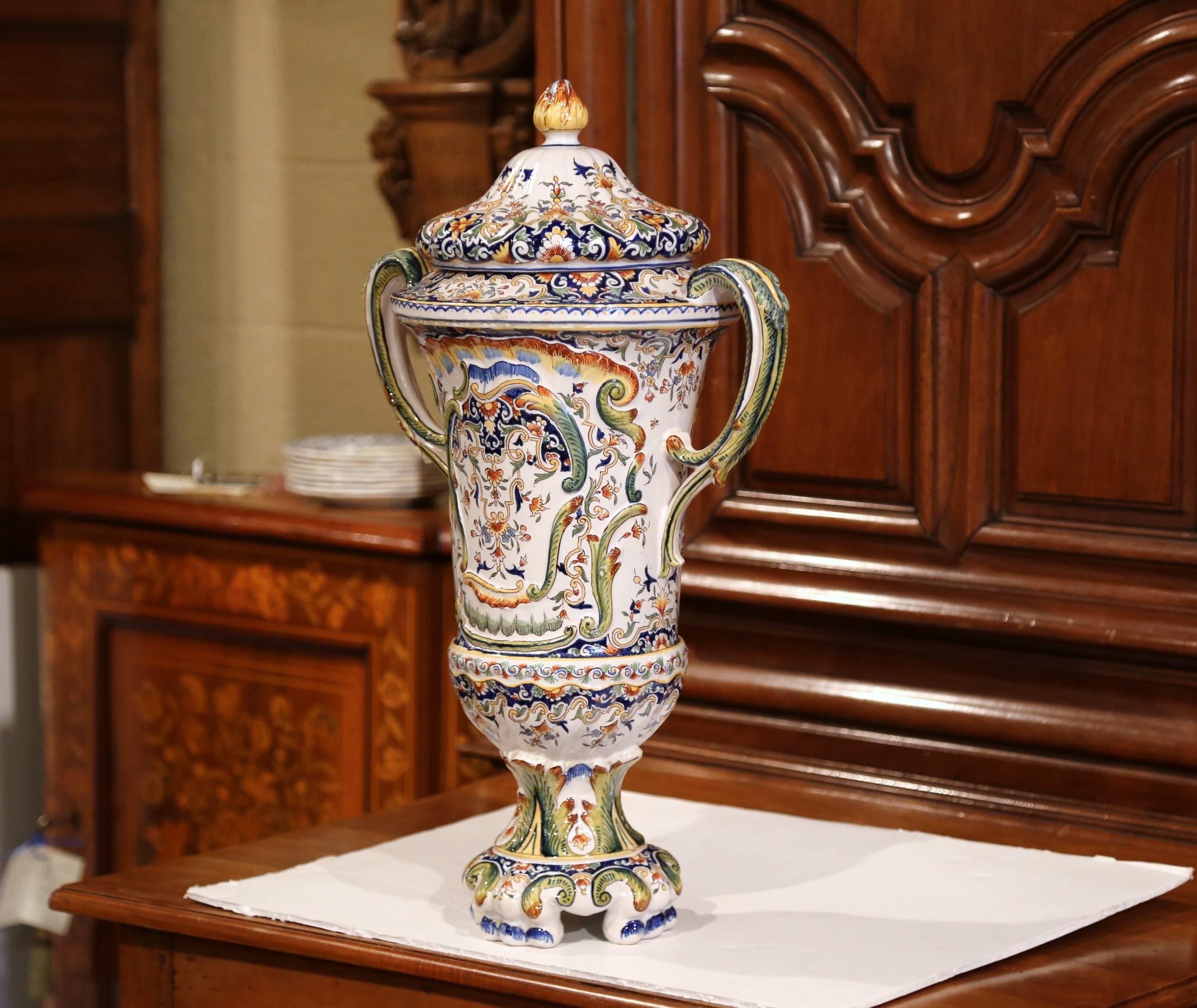 This elegant antique vase was created in Rouen, France circa 1850; the colorful, ceramic vessel stands on small scrolled feet, and features a cone lid with finial at the pediment, embellished with two curved handles with painted leaf motifs in