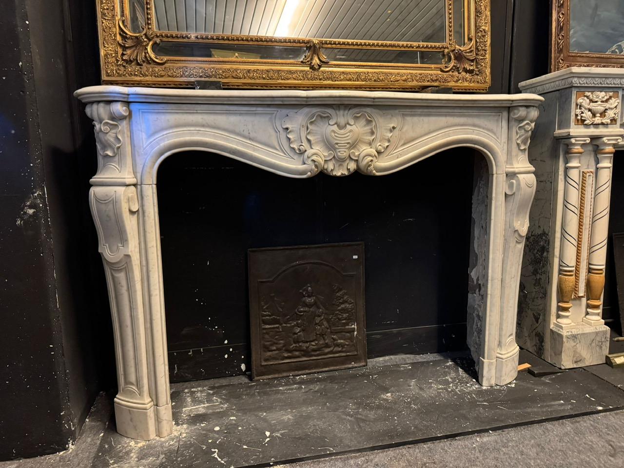 An impressive and beautiful antique fireplace mantel, richly carved in precious white Carrara marble, features a striking and intricate central shell, with small lateral columns and curved legs. Original from the 18th century, it comes from France