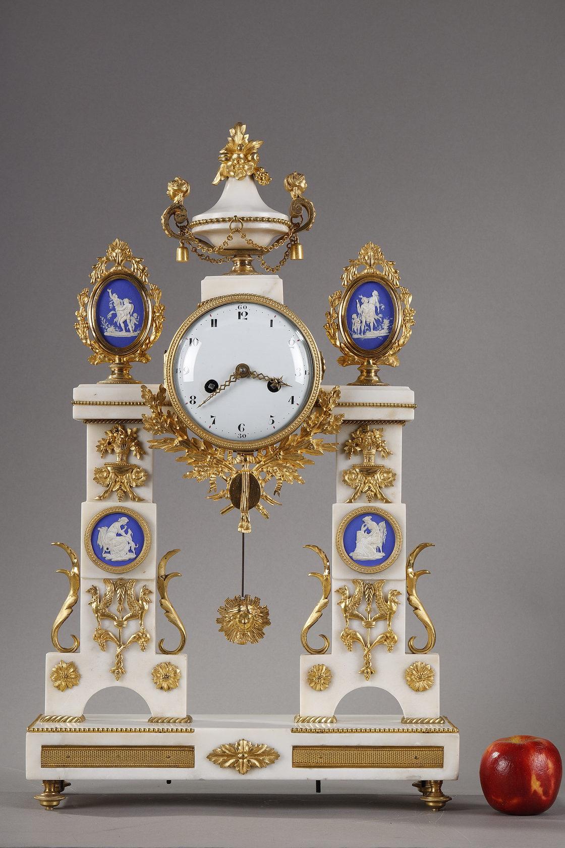 Large Louis XVI period portico clock in white marble, gilt bronze and blue earthenware in the Wedgwood style. The two posts are decorated with chased and gilt bronze motifs representing griffins, acanthus leaves, rosettes and vases overflowing with