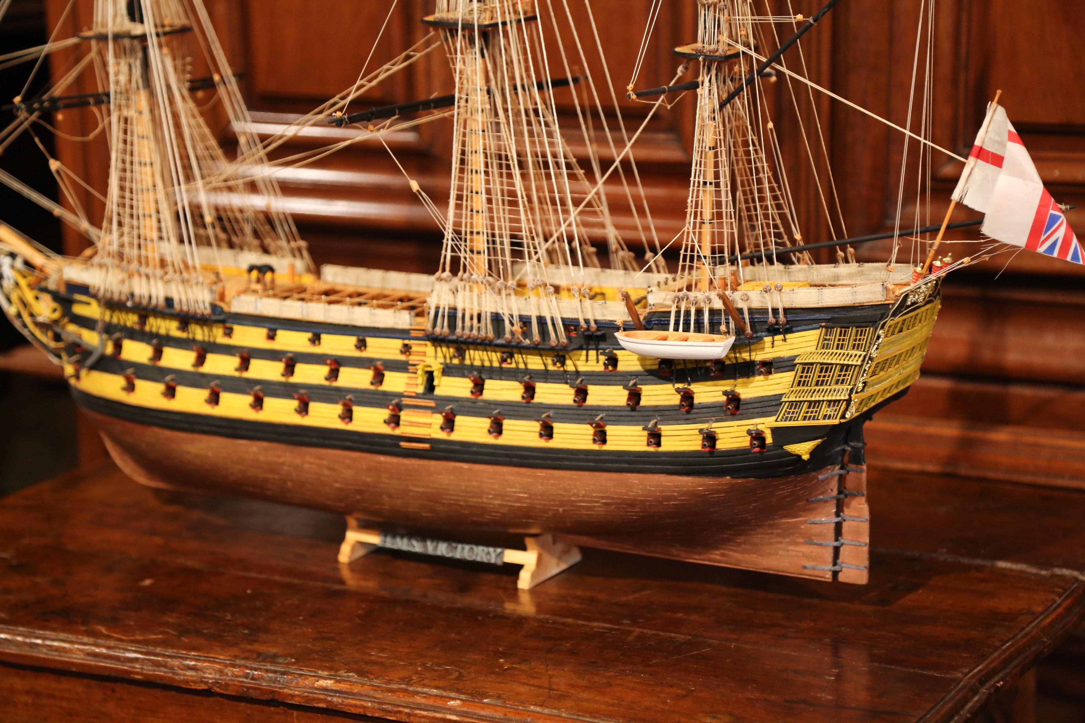 This elegant and majestic ship model was crafted in England, circa 1950. Named H.S.M. Victory, the large naval sailboat sits on a separate wooden base and features authentic and unique details including hundreds of string rigging, spare boats, flags