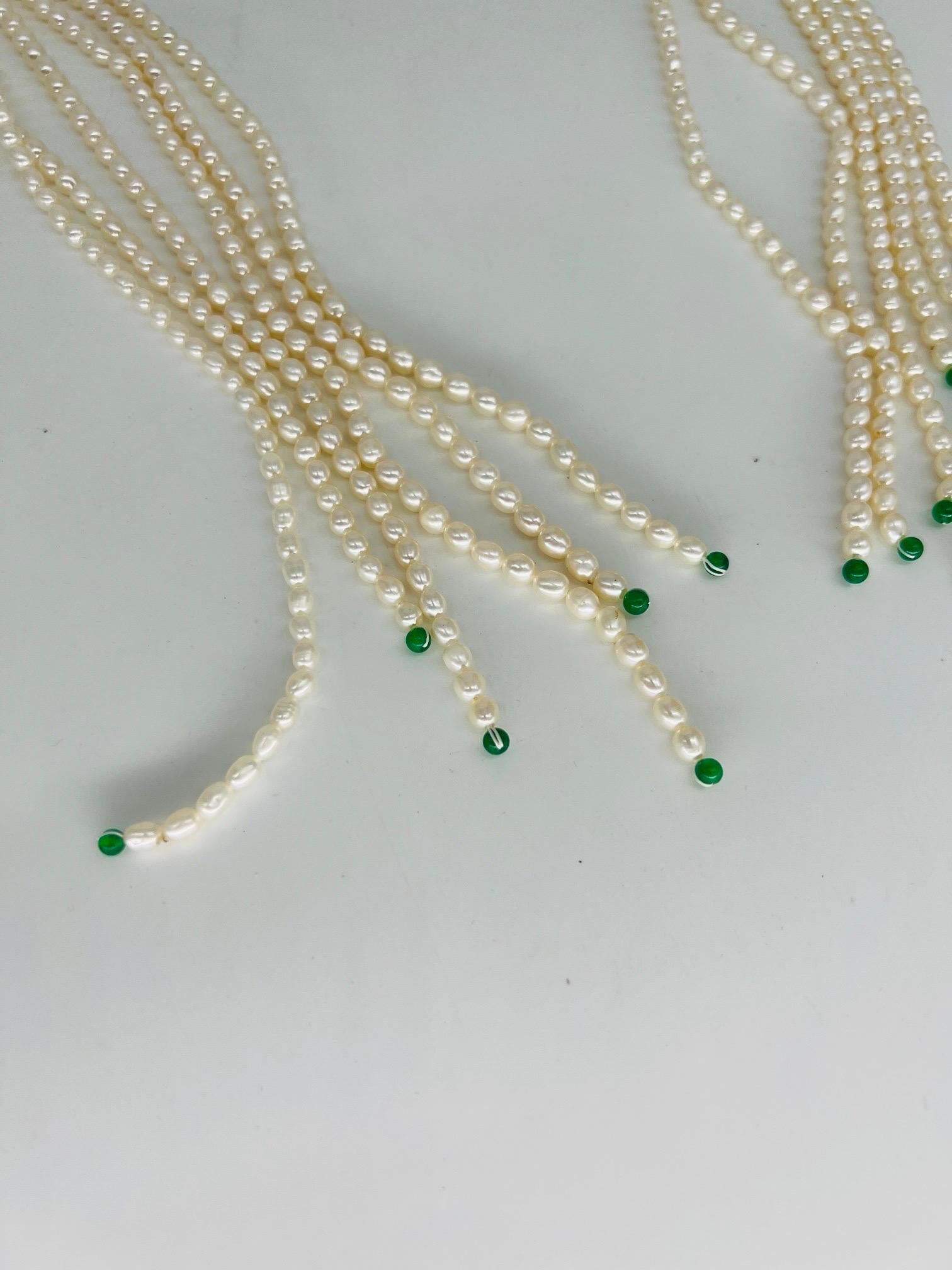 This important necklace features 6 strands of 39.25” string pearls which terminate to tiny green jade rounds. The strands are kept together by a 1.25” w x 1” h green jade necklace “clasp”. The “clasp” adjusts and allows the pearl strands to be set