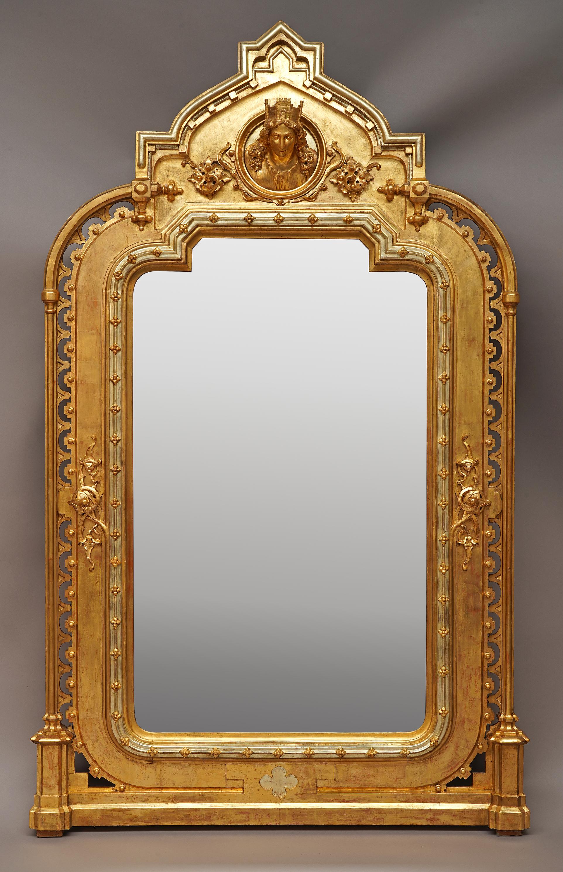 Important miroir en bois sculpté et doré avec deux nuances d'or, de forme architecturale d'inspiration néo-gothique. Bordée de deux colonnes cannelées enfermant des arcs ajourés, reposant sur deux pilastres. Le fronton géométrique est décoré d'un