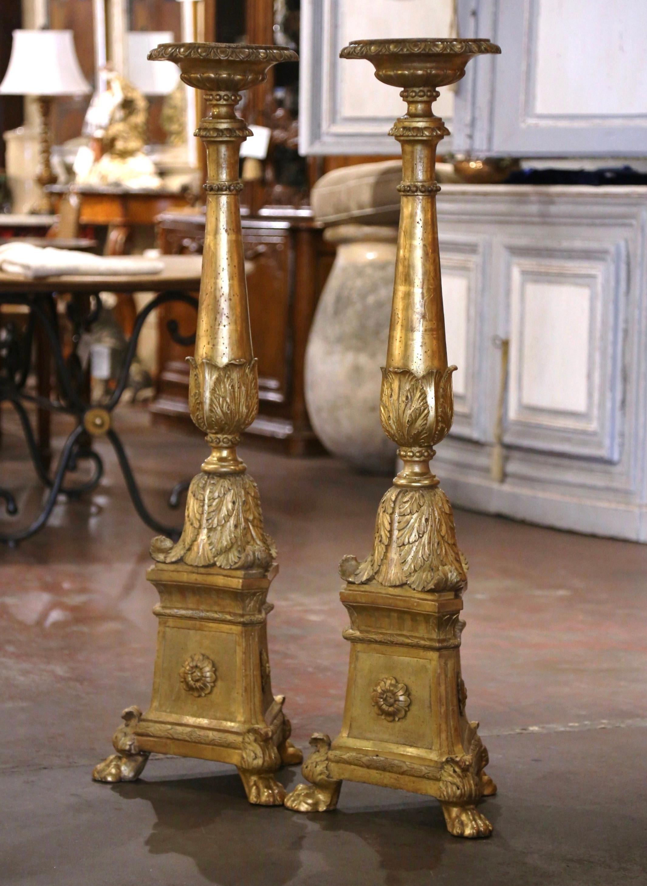 These important antique gilt candlesticks were created in Italy circa 1850. Over 4 feet in height, and standing on three clawed feet over a triangular carved base embellished with relief floral designs and tiered detailing, each tall candlestick