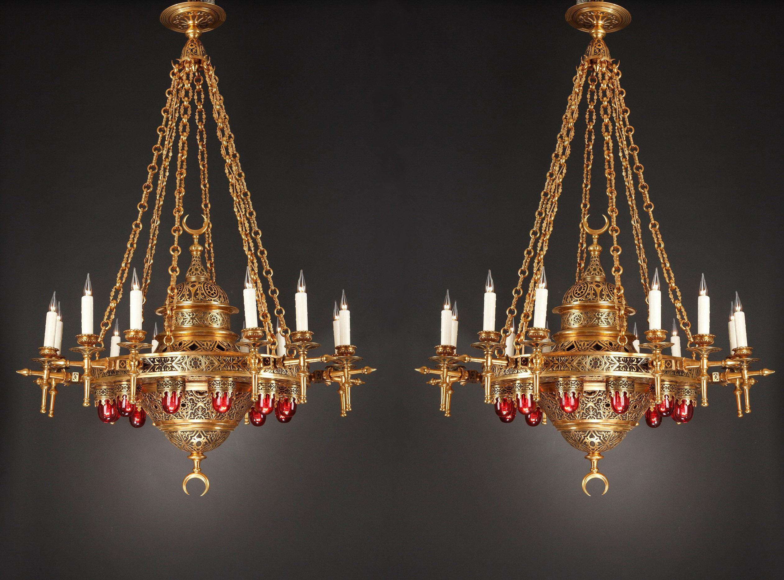 Important pair of Oriental style chandeliers in gilded and patinated bronze suspended by six articulated chains reunited by an elegant roof light. Circular in shape, they have a serie of twelve light-arms around the crown and twelve red verrines