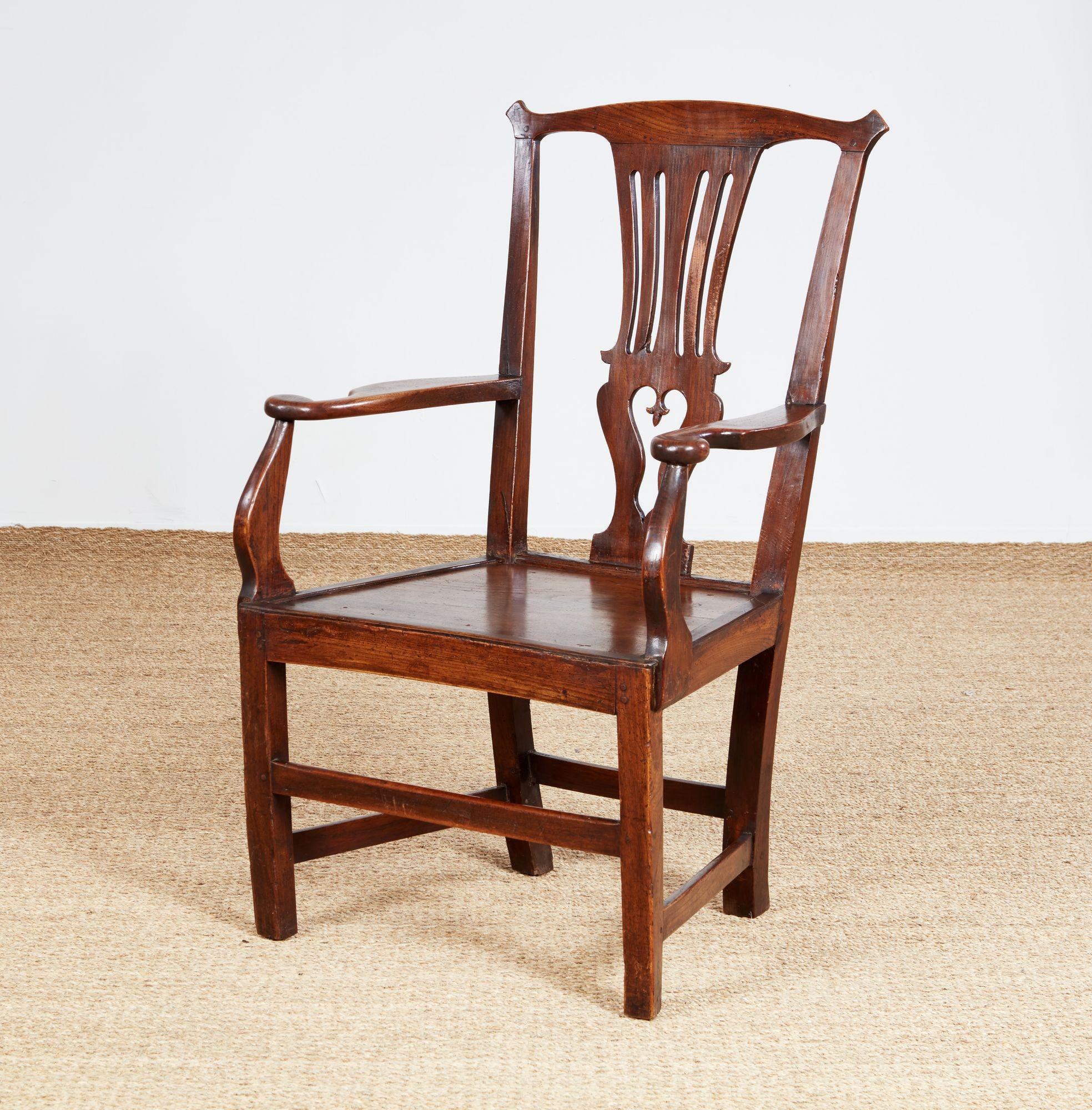 Sillón de roble de generosas dimensiones y porte imponente, con respaldo ligeramente inclinado y barandilla superior ligeramente arqueada con orejas prominentes sobre un respaldo calado con flores de lis talladas en forma de gota, reposabrazos