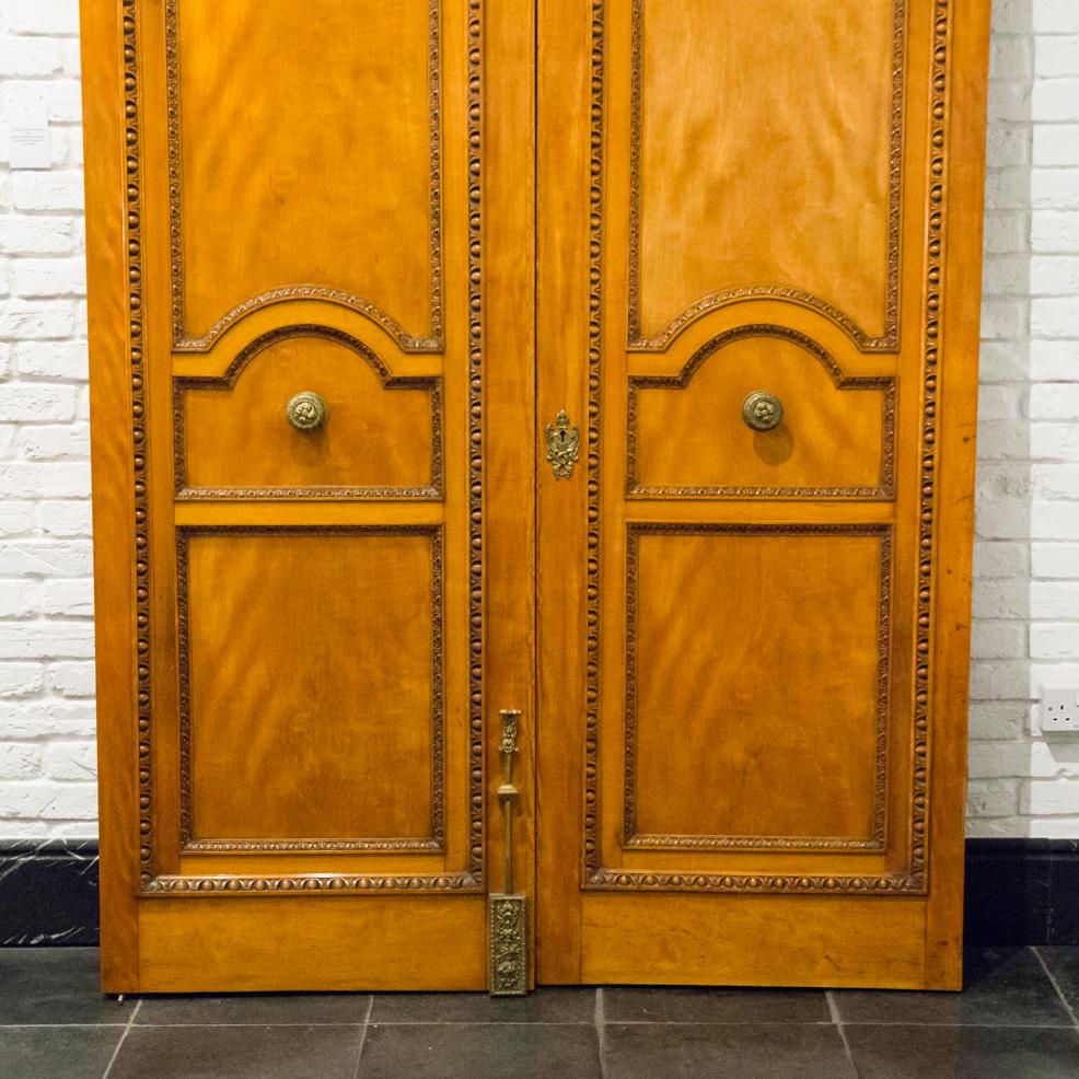 A pair of monumental solid satinwood doors with classical egg and dart carved decoration and fine quality brass door furniture.

Brass door pulls marked CHING (Comyn Ching of London).

Full set of brass acorn finial hinges included.

Left