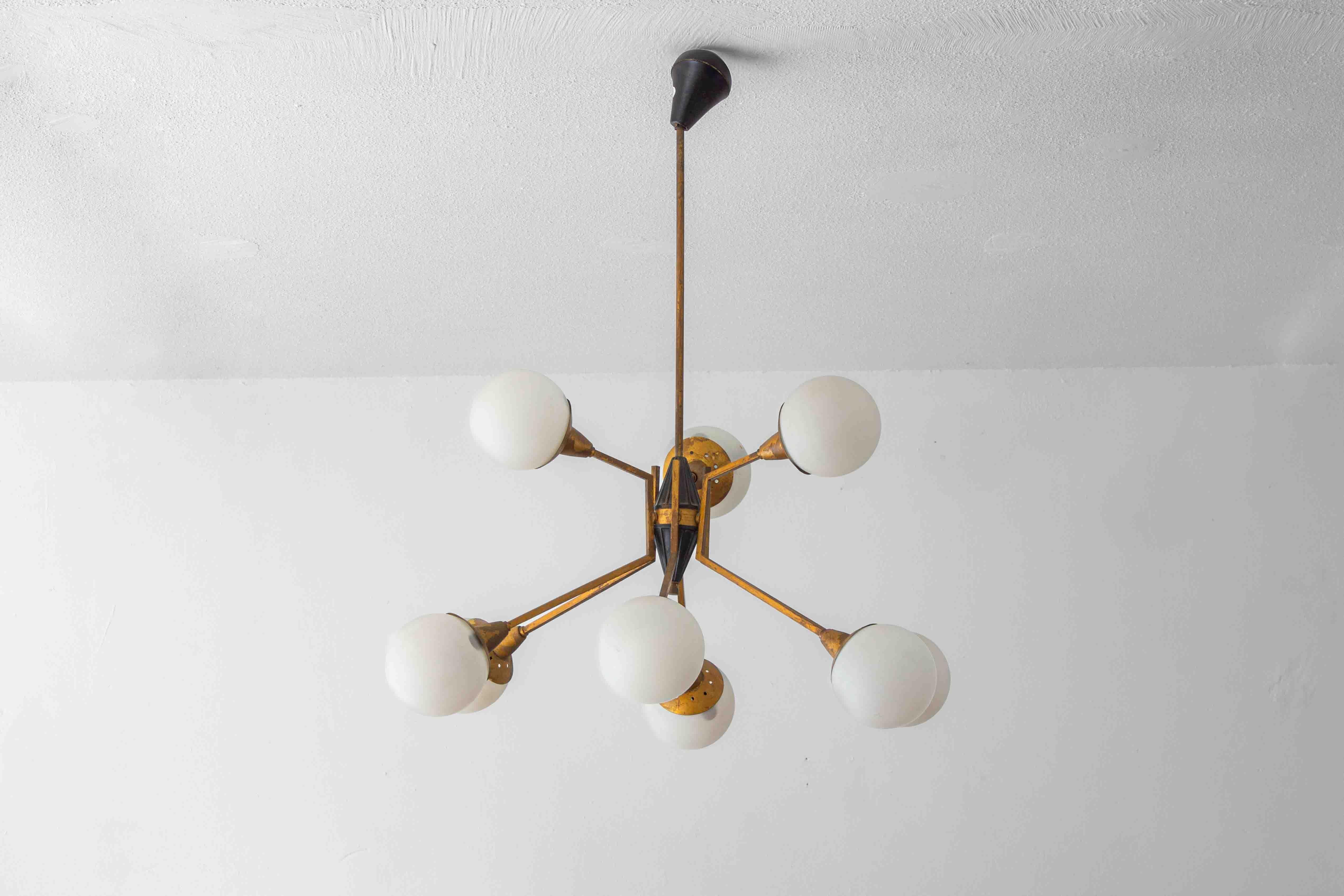 Lustre italien du milieu du siècle, composé de neuf bras disposés de façon radiale. Chaque bras porte un globe en verre opalin, relié par des tubes en laiton à la patine chaude. Le corps central est en laiton avec des détails en émail noir.

Le