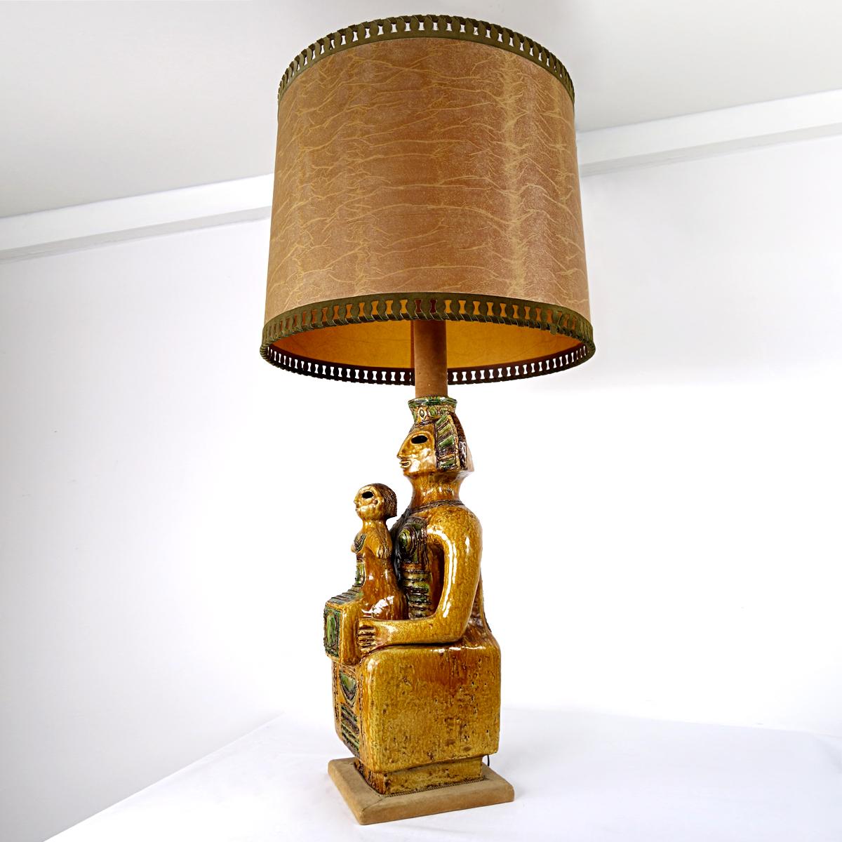 This eye catching floor or table lamp in Mayan style truly is an impressive piece.
Its foot is a large ceramic statue of a queen and her child, or a king if you will with his successor, in brown and green shades. 
The lower part of the 