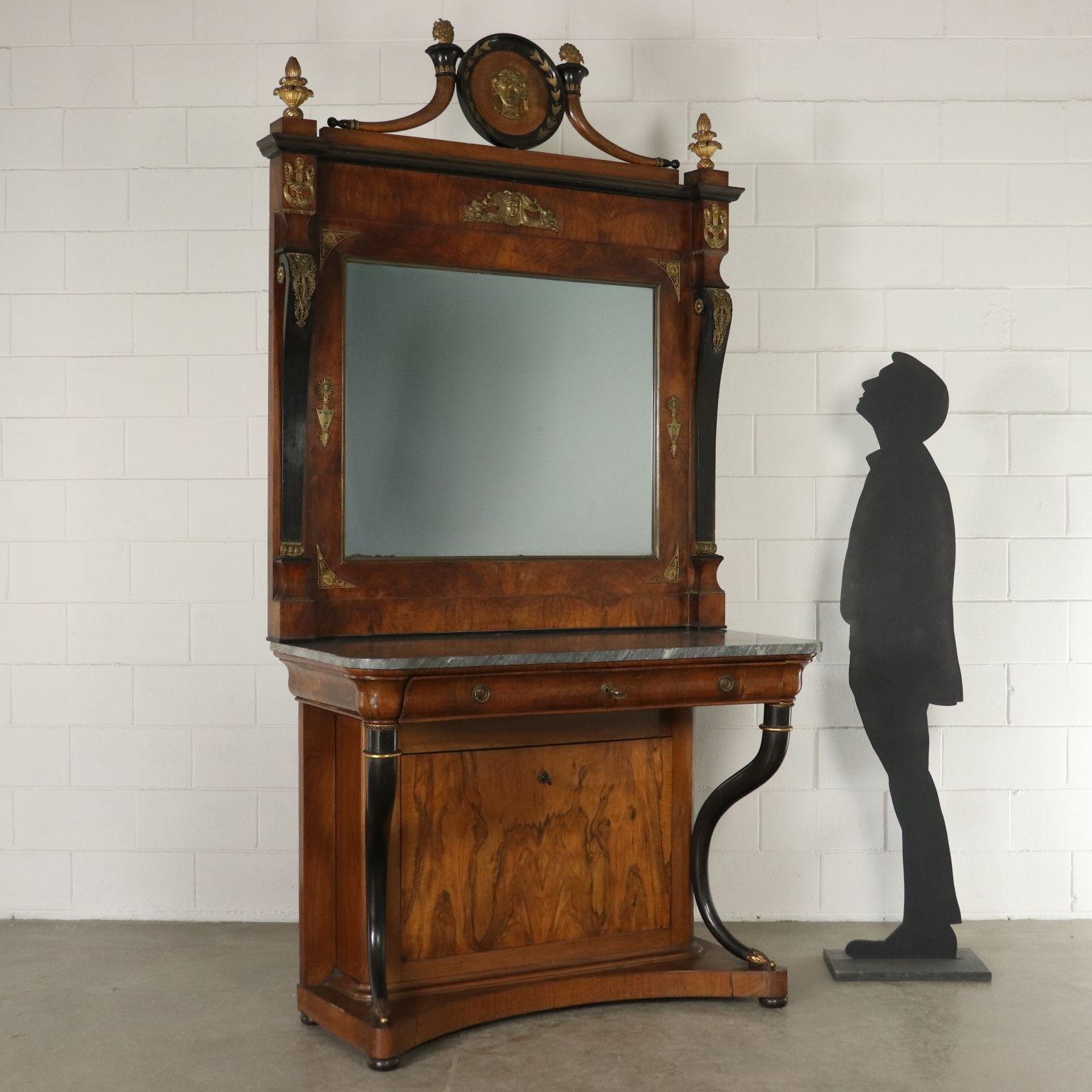 An impressive console with mirror. Ebonized cornucopias with gilded carvings. Shaped drawer under the top and drop-leaf door hiding a compartment. Marble top. Elegant coeval mirror with ebonized pilaster strips, frieze with medal and cornucopias