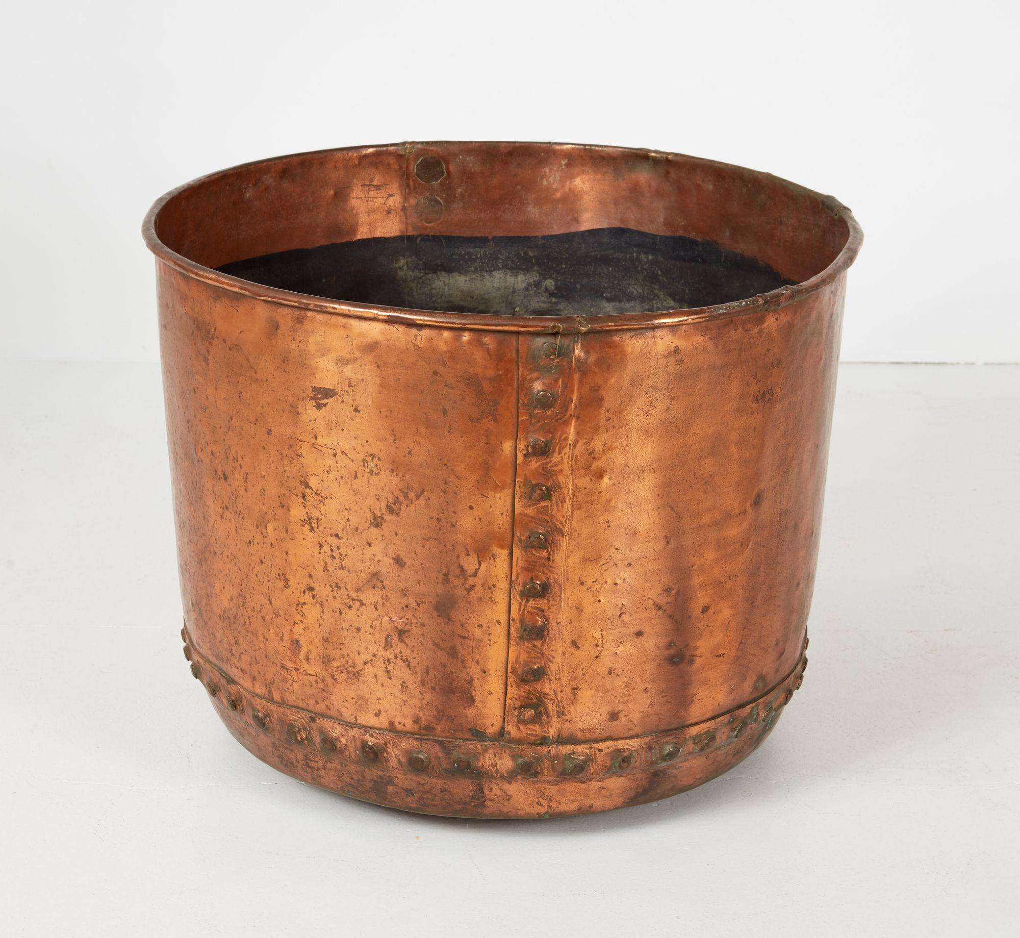 A large copper vessel, with rolled rim and gently canted sides, possessing functional and decorative riveted seams and being of particularly impressive circumference. Useful as a large log bin, an outdoor feature planter filled with tulip bulbs and