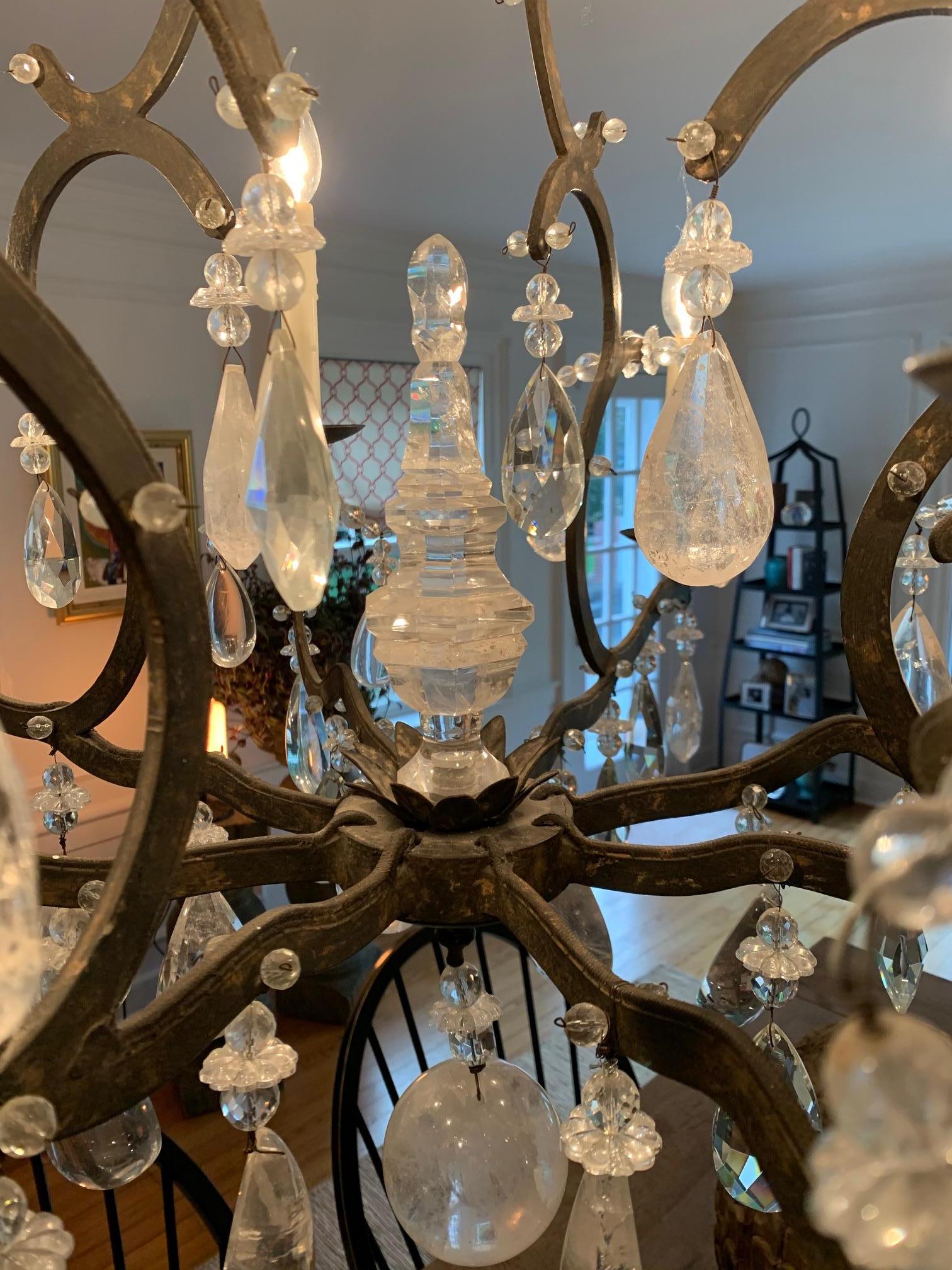 A very large 8 arm wrought iron and crystal chandelier having chunky tear dropped aged crystals dripping all over it, a central crystal finial and large round ball at the bottom. The iron is a dark brown and the overall effect is rustic glamour.