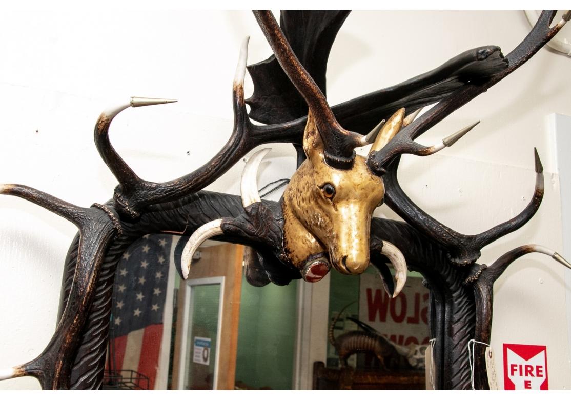 Impressive figural stag head mirror garnished with faux stag and moose antlers atop a carved rectangular wooden frame. The top with central carved stag head with gilt painted finish and a polished red stone encased in silver tone metal frame resting