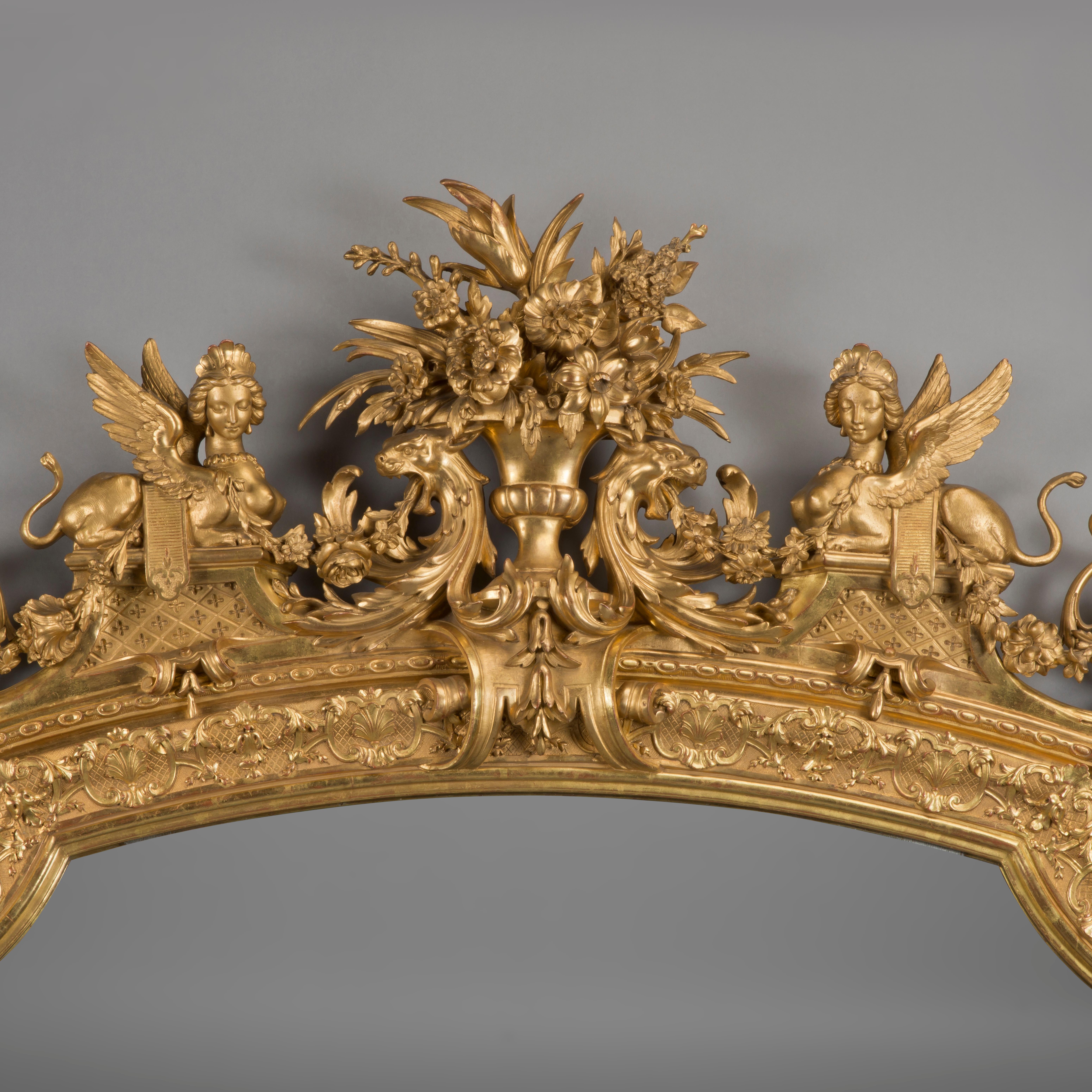 A large and impressive Louis XVI style carved giltwood and gesso mirror.

At over 100 inches tall (260 cm) this impressive mirror is of large size and finely carved with a figural cresting rail centred by a vase issuing flowers flanked by