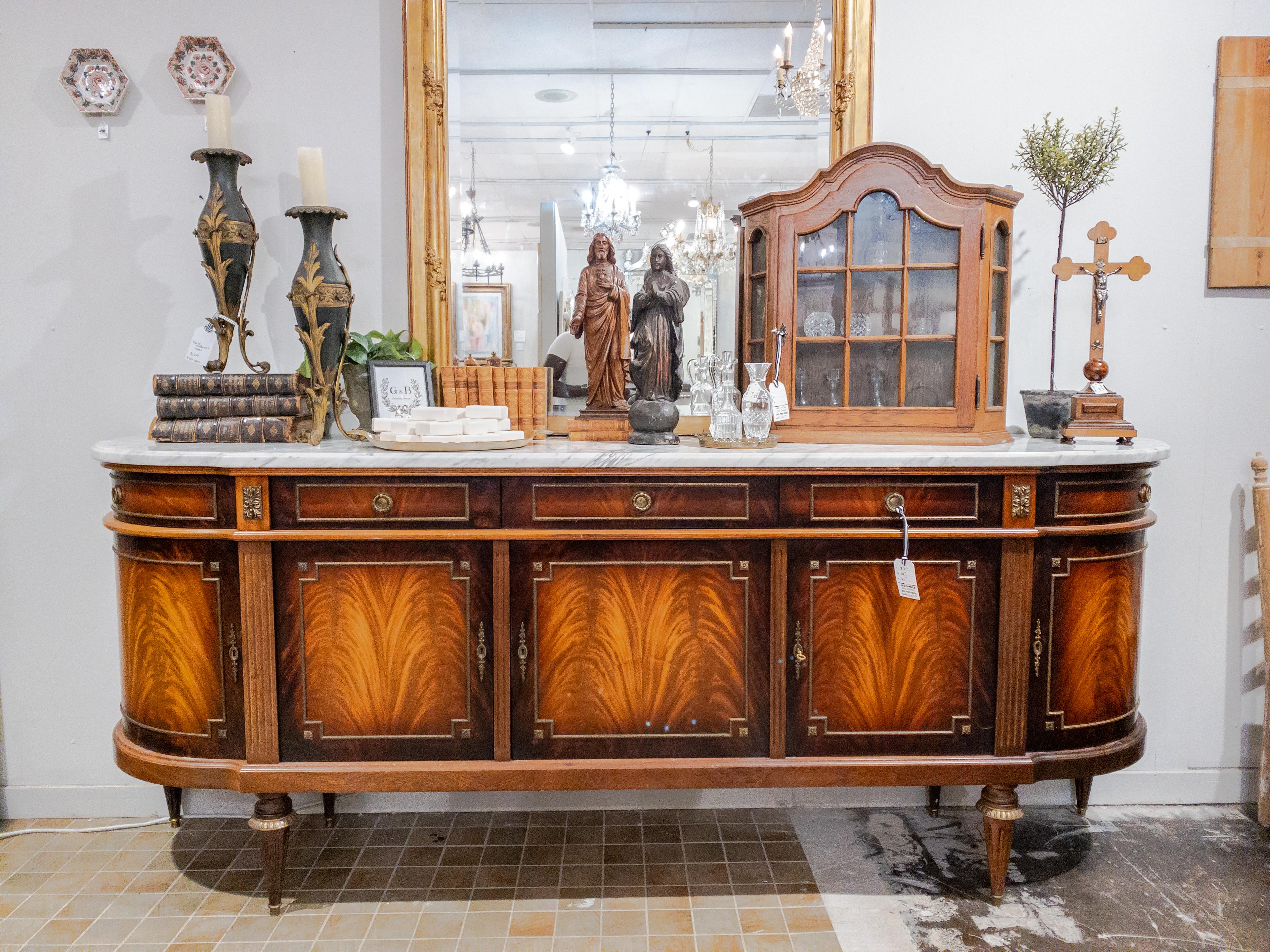 The Impressive Louis XVI Style Marble and Mahogany Buffet is a stunning piece of furniture designed in the classical Louis XVI style, known for its elegance and refinement. The buffet features flame-grained mahogany doors, showcasing the exquisite