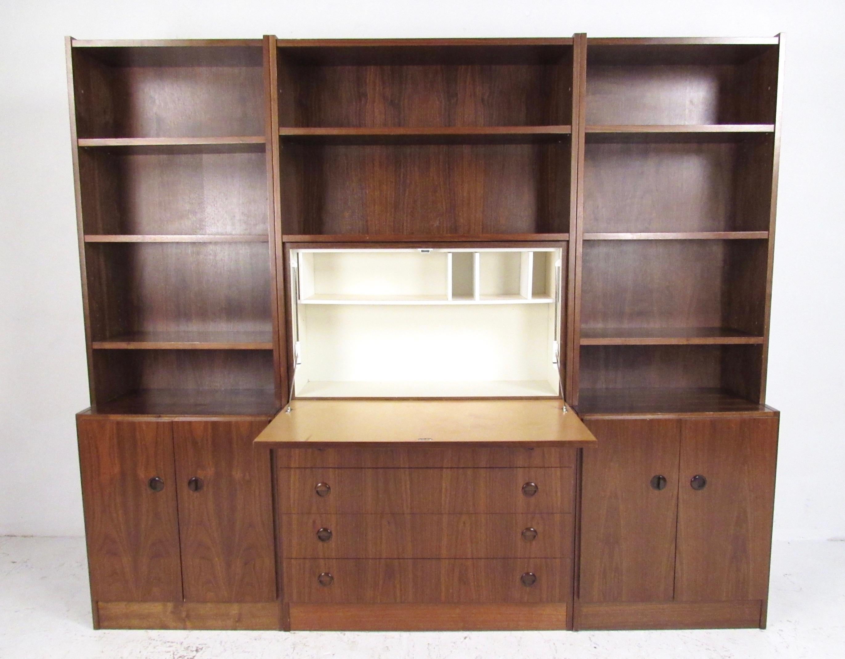 This stylish seven foot wide bookcase features vintage walnut finish, spacious cabinet and drawer storage, and unique drop front work desk or bar area. Adjustable shelves and quality midcentury construction, this piece was originally produced in