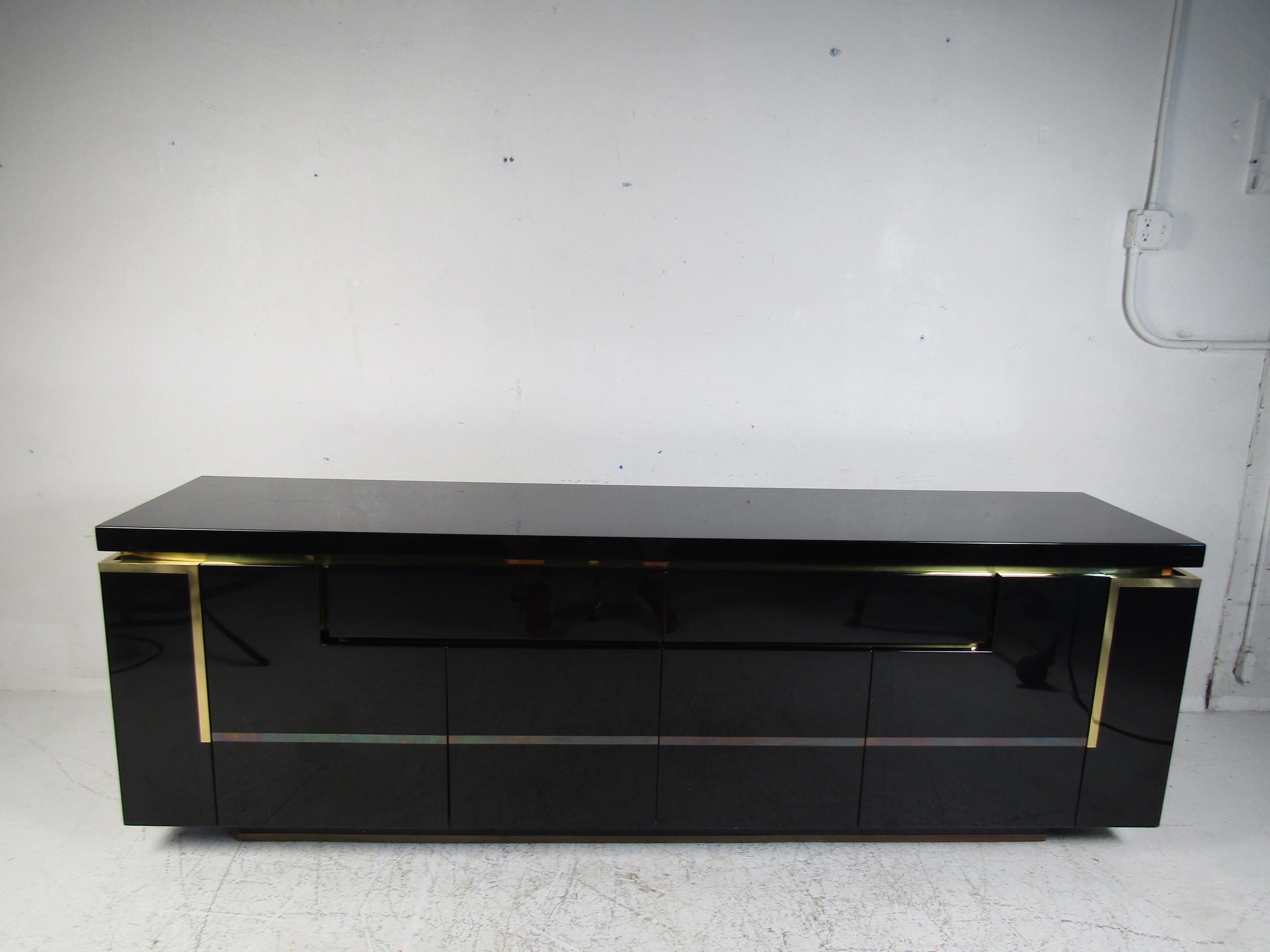 This stunning modern sideboard offers plenty of room for storage within its many compartments. The unique design appears to have interlocking cabinets with the two drawers. The glossy black finish and brass trim add to the allure. A finished back