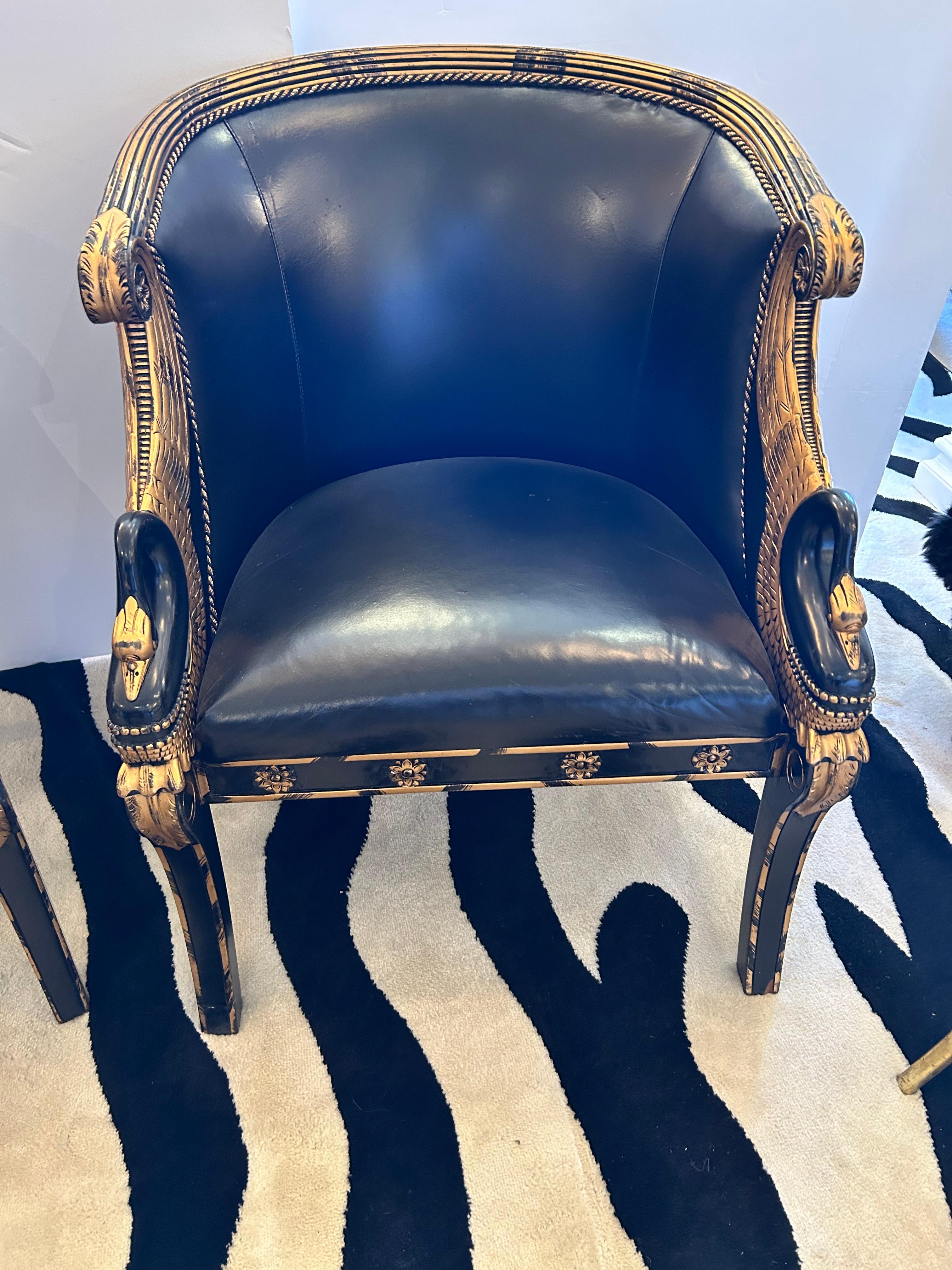 Impressive pair of Empire style black and gilded wood club chairs having black leather upholstery, curved backs and arms in the form of swans resting on saber shaped legs.  The backs are upholstered in a complimentary striped fabric.
