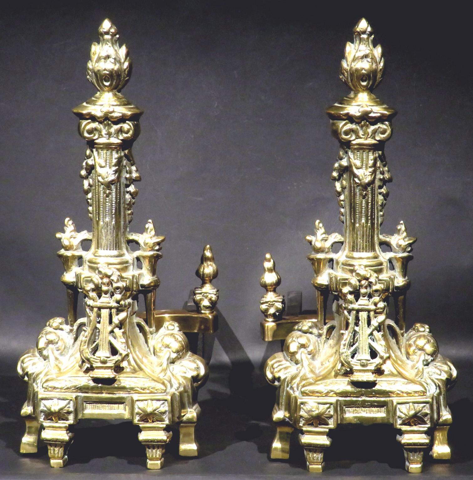 A large and impressive pair of Louis XVI style gilt bronze chenets showing applied lyre shaped elements fronting torchere shaped columns with flame shaped finials.