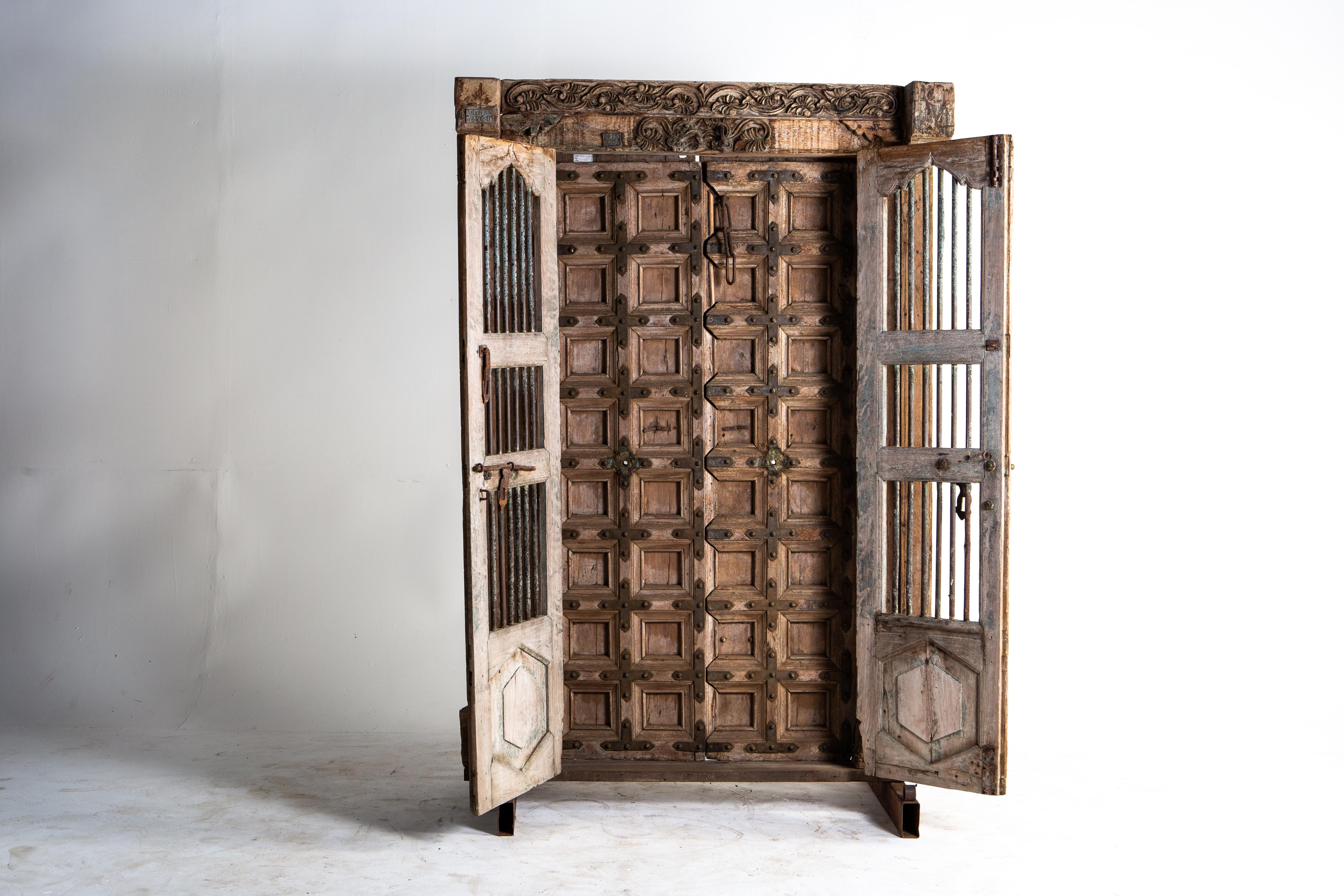 This complete entryway is handsomely relief carved; the frame features intricate floral motifs and geometric patterns throughout. The panelled doors, with metal studs and hardware, open inwardly and can be secured with a chain and pin lock. The