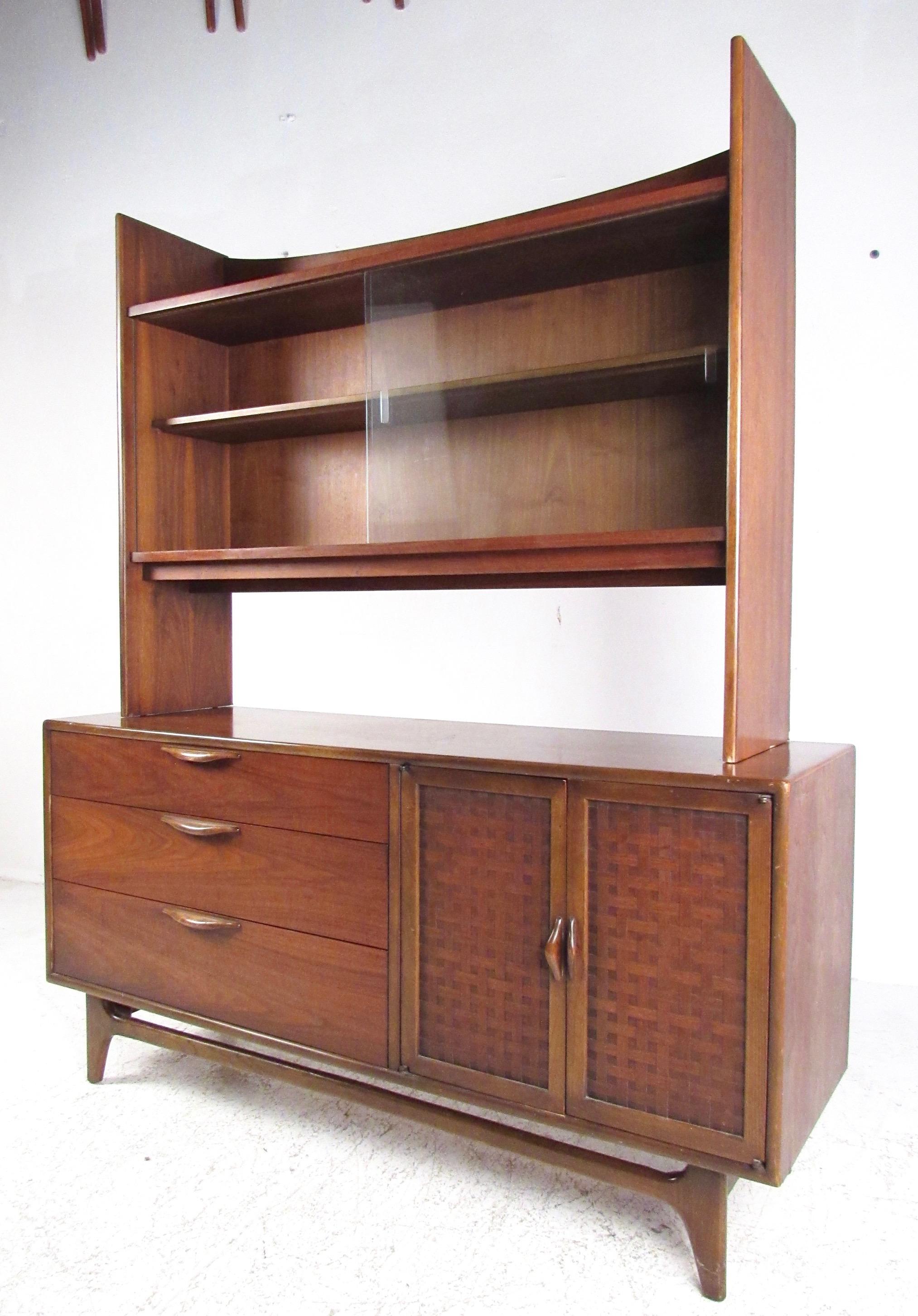 This Mid-Century Modern sideboard features iconic Warren Church design for Lane, including carved wood handles and basket woven details. The upper display cupboard features shapely design and sliding glass doors, adding to the impressive vintage