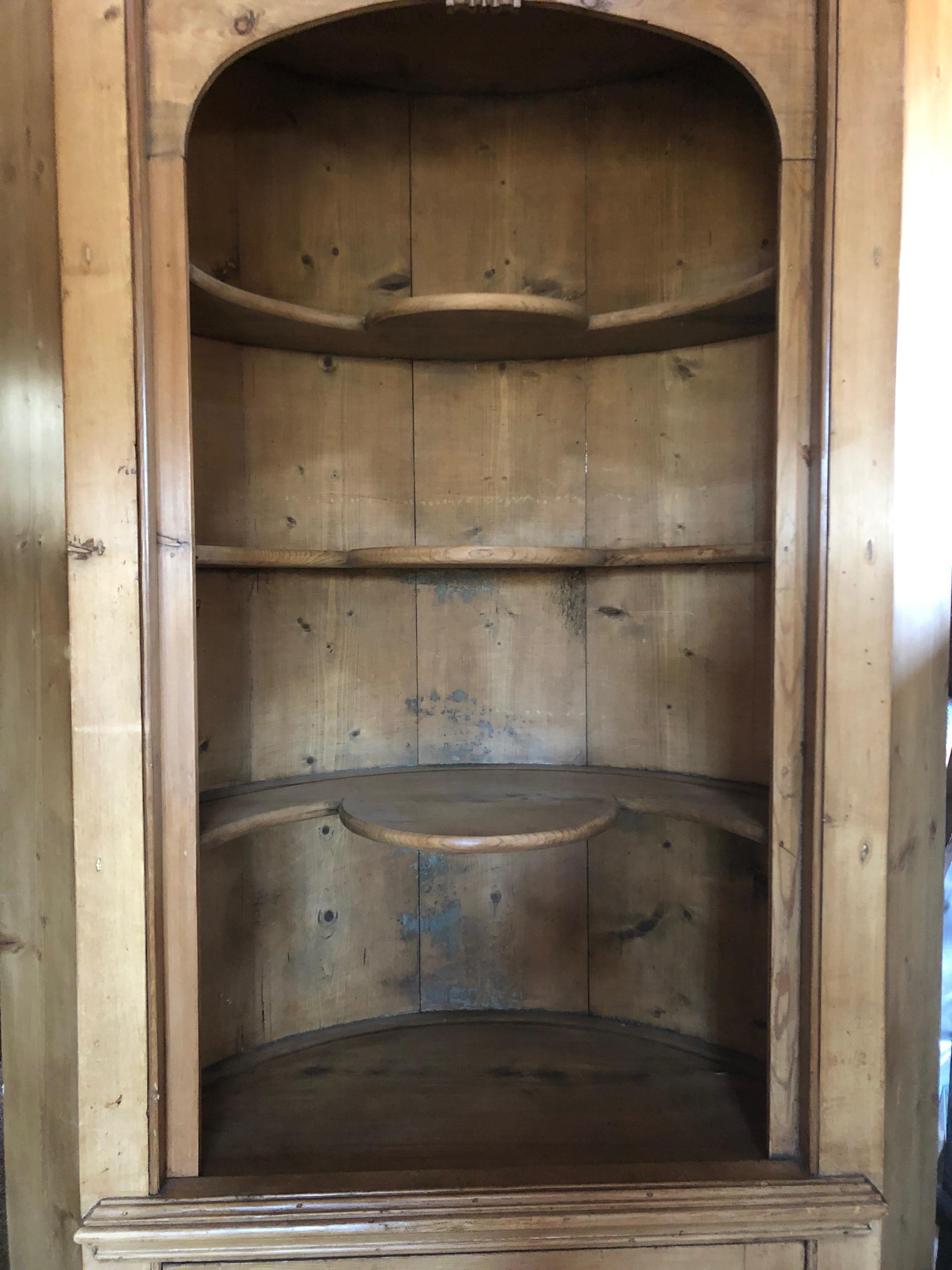 Very tall impressive natural unfinished pine corner cupboard having a carved wood cornice, 3 curvy interior exposed shelves, a carved wood door that opens to reveal storage inside, and a barrel shaped back measuring 49 inches round.
18 inches deep