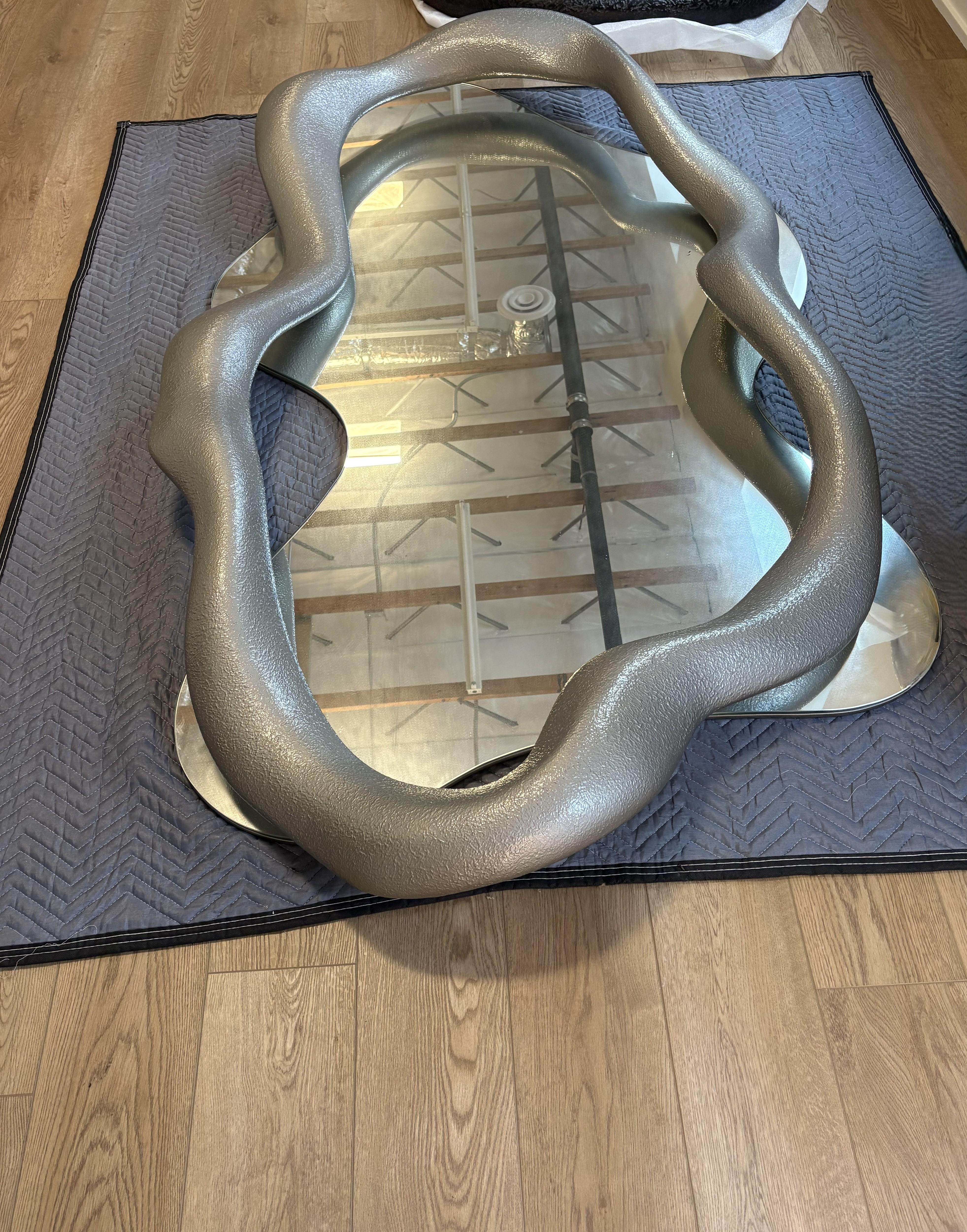 Ein auffälliger, skulpturaler Wandspiegel mit einem wellenförmigen, organischen Rahmen, der mit einer strukturierten Zinnpatina überzogen ist, ist jetzt auf Lager. Die fließende Silhouette des Spiegels lässt die Grenze zwischen Kunst und Funktion